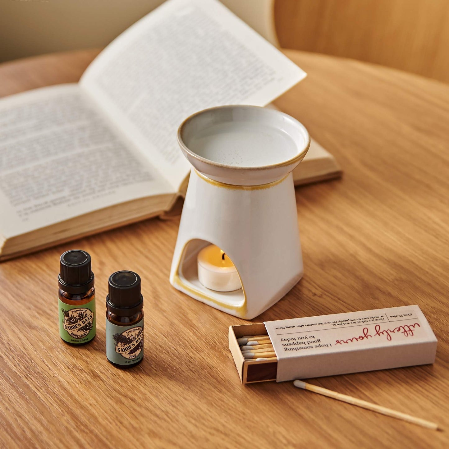나무 테이블 위 펼쳐진 책과 성냥갑 옆에 놓인 화이트 세라믹 옐로우 오일버너와 불이 켜진 티라이트 캔들, 앞에 세워둔 가든데이즈 에센셜오일 병 두 개가 보이는 독서 연출 사진