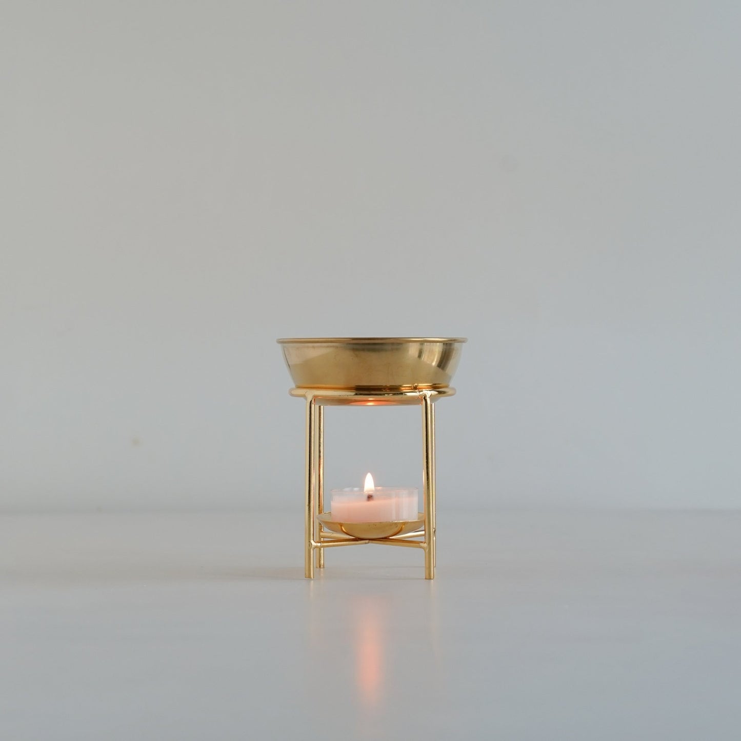 연한 배경 앞 단독으로 놓인 골드 스탠드 오일버너와 안에서 은은하게 빛나는 티라이트 캔들을 정면에서 담은 제품 사진