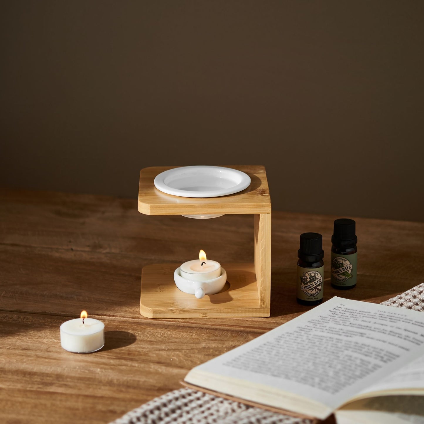 우드 테이블 위에 내추럴 우드 오일버너와 그 앞의 티라이트 캔들, 옆에 놓인 가든데이즈 에센셜오일 3종과 펼쳐진 책이 함께 보이는 선물세트 사용 연출 사진