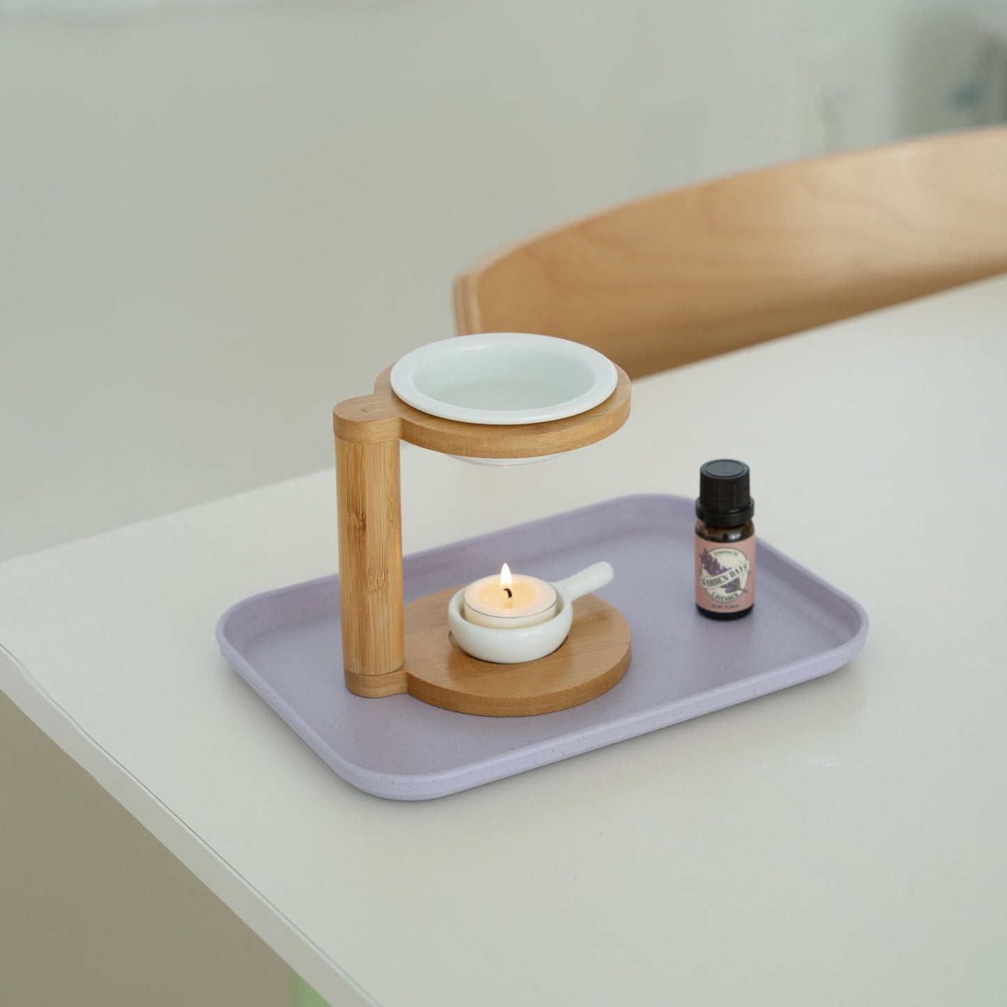 보라색 트레이 위에 놓인 우드 오일버너와 불이 켜진 티라이트 캔들, 옆에 세워둔 가든데이즈 에센셜오일 병이 보이는 테이블 연출 사진