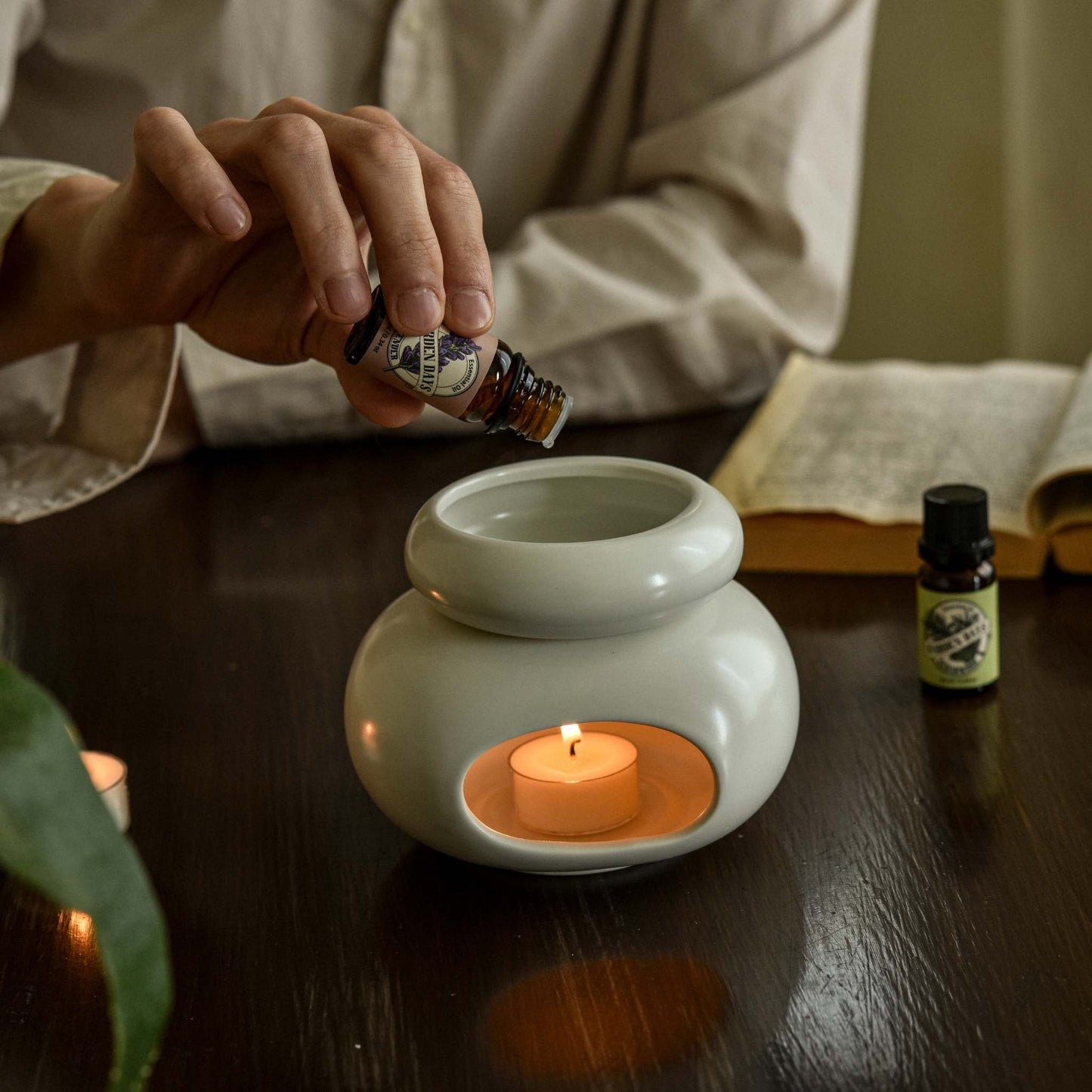 테이블 위 조약돌 도자기 오일버너 안에서 티라이트 캔들이 켜져 있고, 옆에 가든데이즈 천연 100% 에센셜오일 병들이 놓인 아로마 테라피 연출 사진