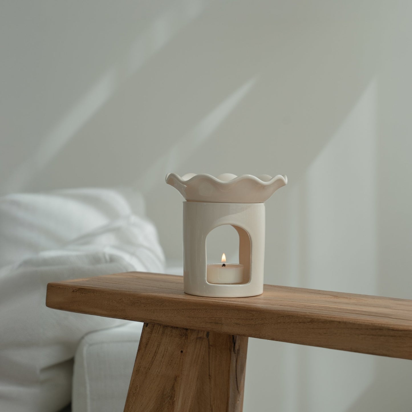 창가 쪽 나무 벤치 끝에 놓인 웨이브 모양 화이트 세라믹 오일버너와 안에서 켜진 티라이트 캔들을 옆에서 담은 연출 사진