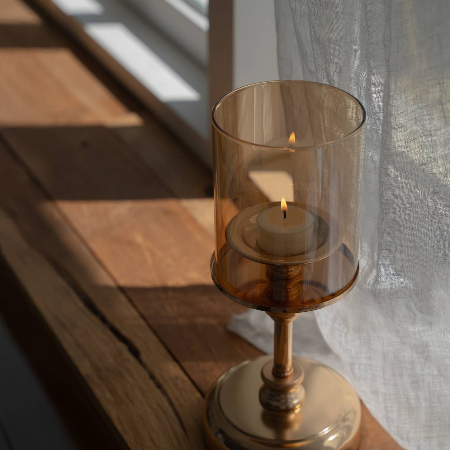 창가 목재 선반 위에 놓인 브라운 톤 유리 갓의 골드 스탠드 캔들 홀더와 안에서 밝게 켜진 티라이트 캔들을 가까이에서 담은 클로즈업 사진