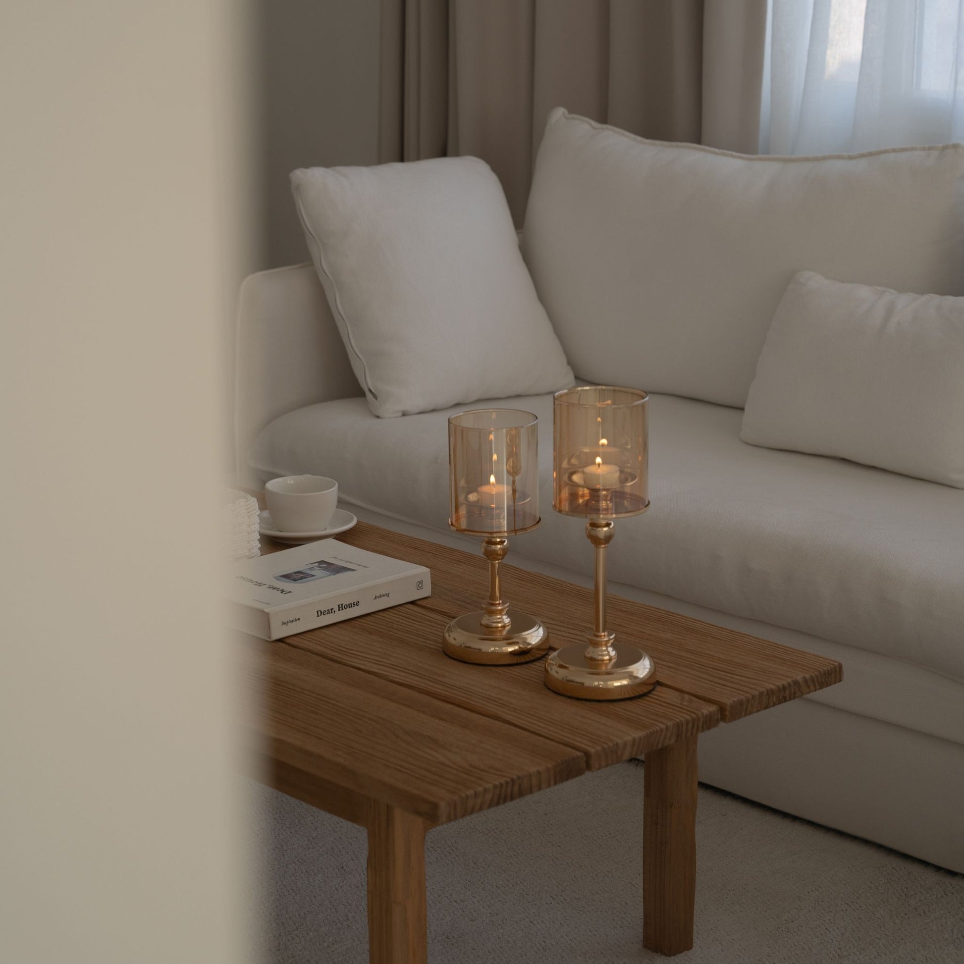 소파 옆 나무 티테이블 위에 같은 디자인의 골드 스탠드 유리 캔들 홀더 두 개와 켜진 티라이트 캔들, 책이 함께 놓인 거실 연출 사진