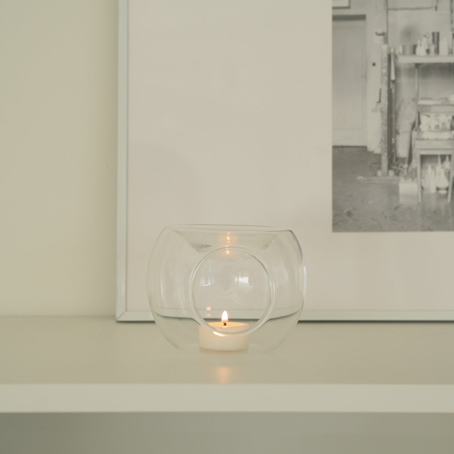 화이트 선반 위 액자와 오브제 앞에 단독으로 놓인 투명 유리 티라이트 홀더와 은은한 불빛의 티라이트 캔들이 보이는 미니멀 인테리어 사진