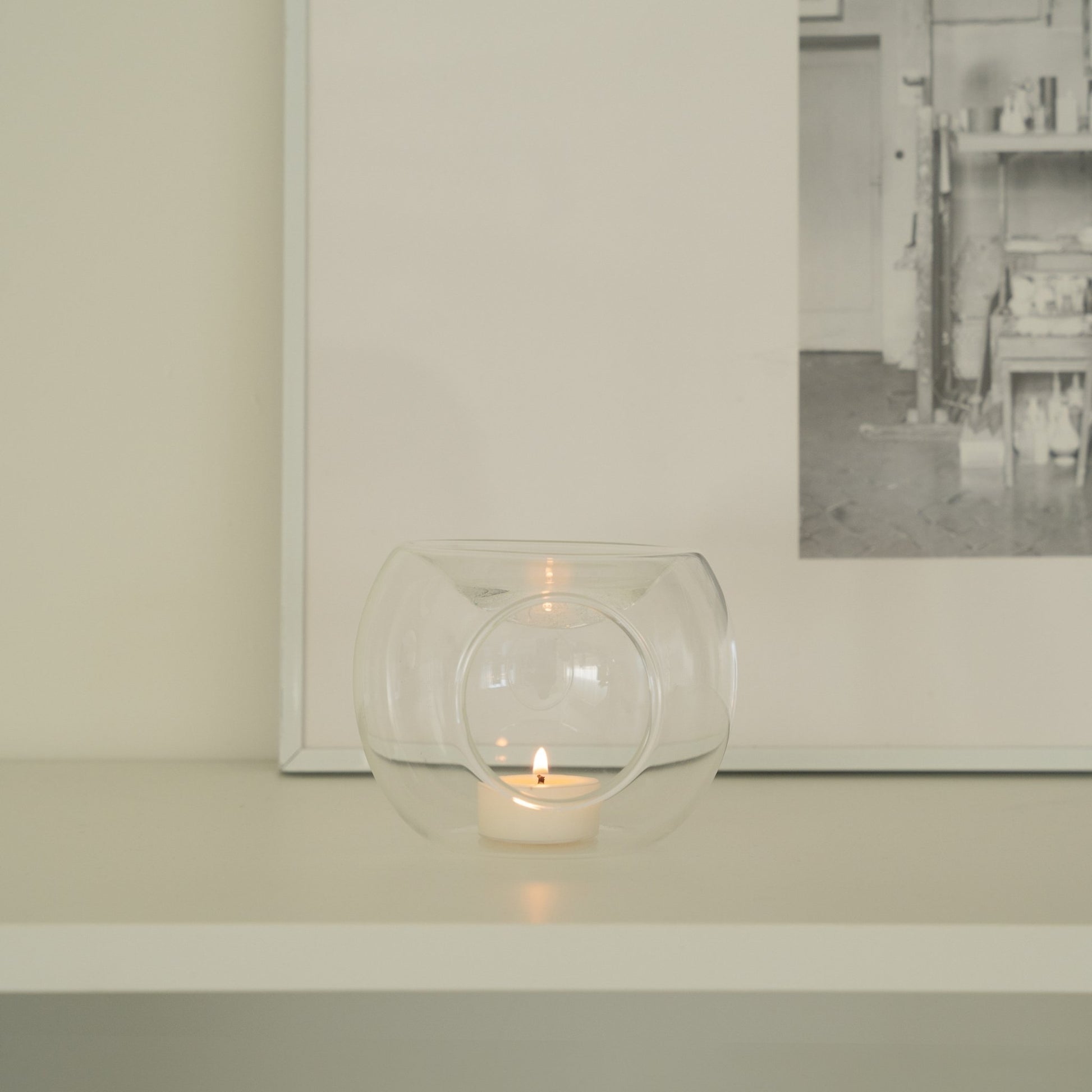 화이트 선반 위 액자와 오브제 앞에 단독으로 놓인 투명 유리 티라이트 홀더와 은은한 불빛의 티라이트 캔들이 보이는 미니멀 인테리어 사진