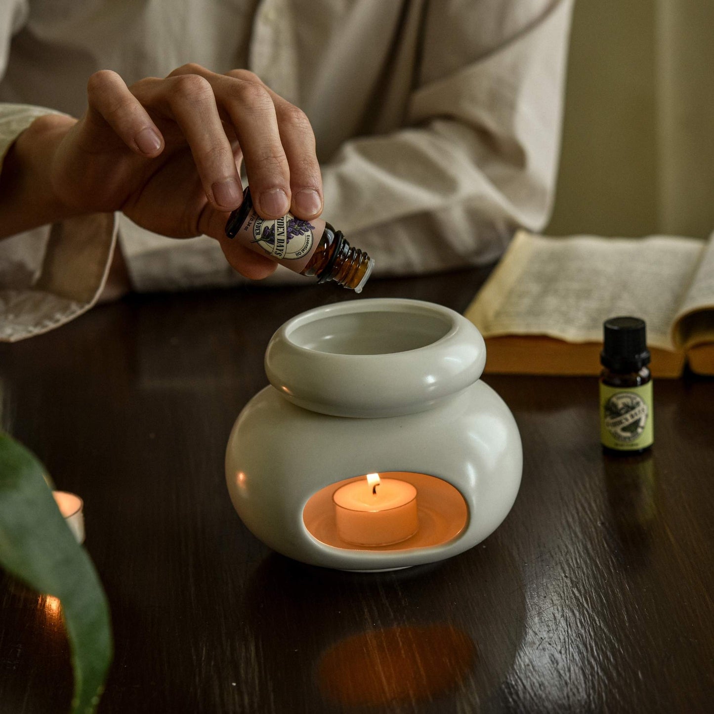 책상 위에 올려둔 조약돌 도자기 오일버너 안 티라이트 캔들 위로 에센셜오일을 떨어뜨리는 손과 옆에 놓인 가든데이즈 에센셜오일 병이 보이는 사용 연출 사진