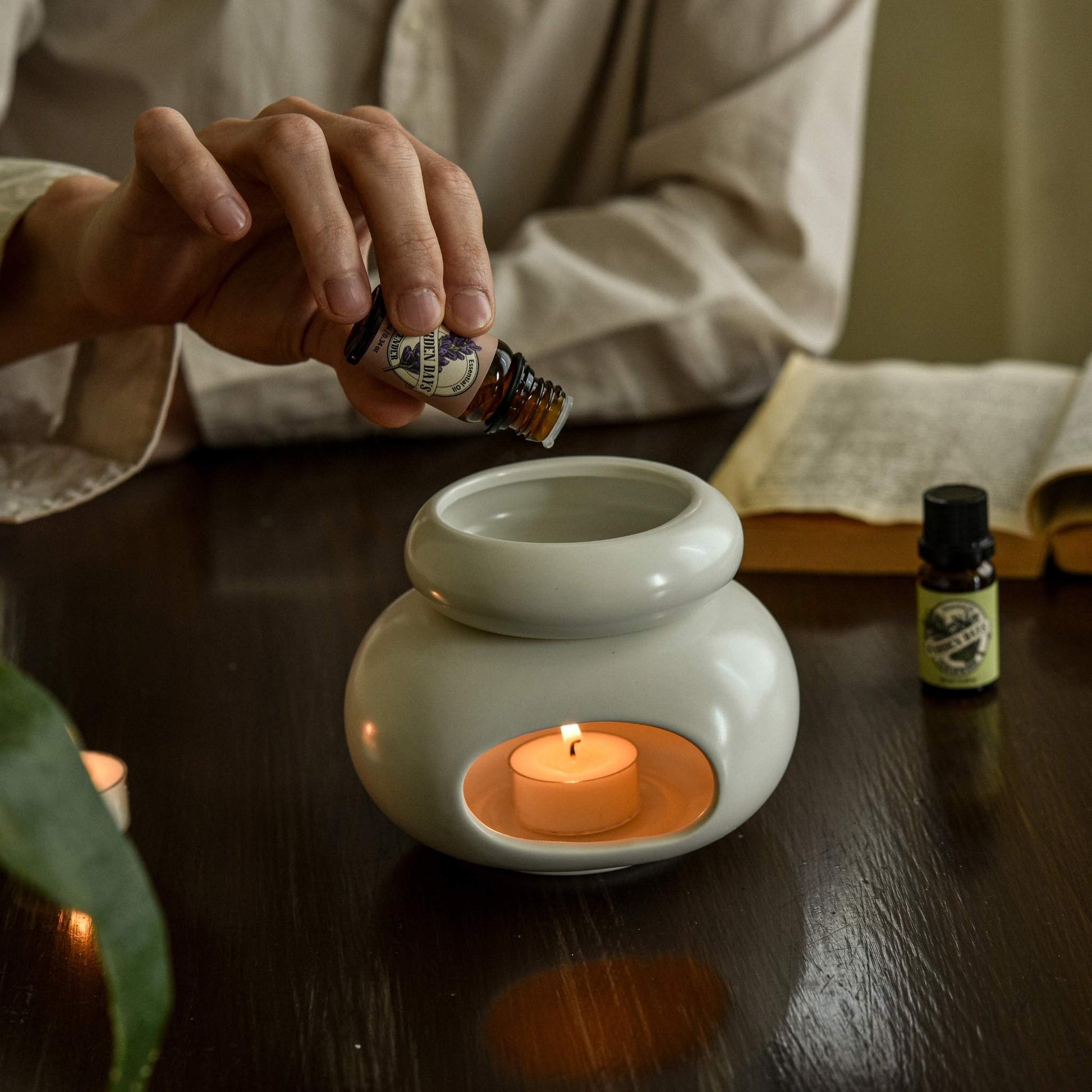 책상 위에 올려둔 조약돌 도자기 오일버너 안 티라이트 캔들 위로 에센셜오일을 떨어뜨리는 손과 옆에 놓인 가든데이즈 에센셜오일 병이 보이는 사용 연출 사진