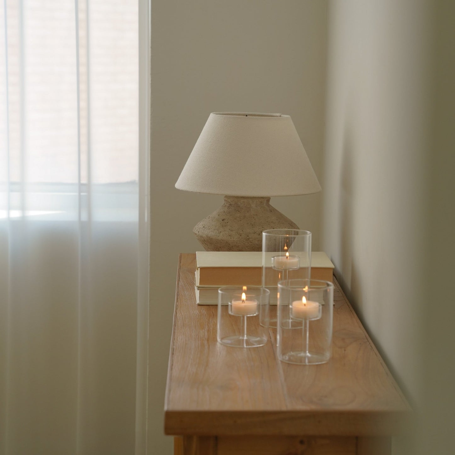 창가 쪽 콘솔 테이블 위 스탠드 조명과 책 옆에 올려진 투명 유리 실린더 캔들 홀더 세트와 은은한 불빛의 티라이트 캔들이 보이는 인테리어 연출 사진