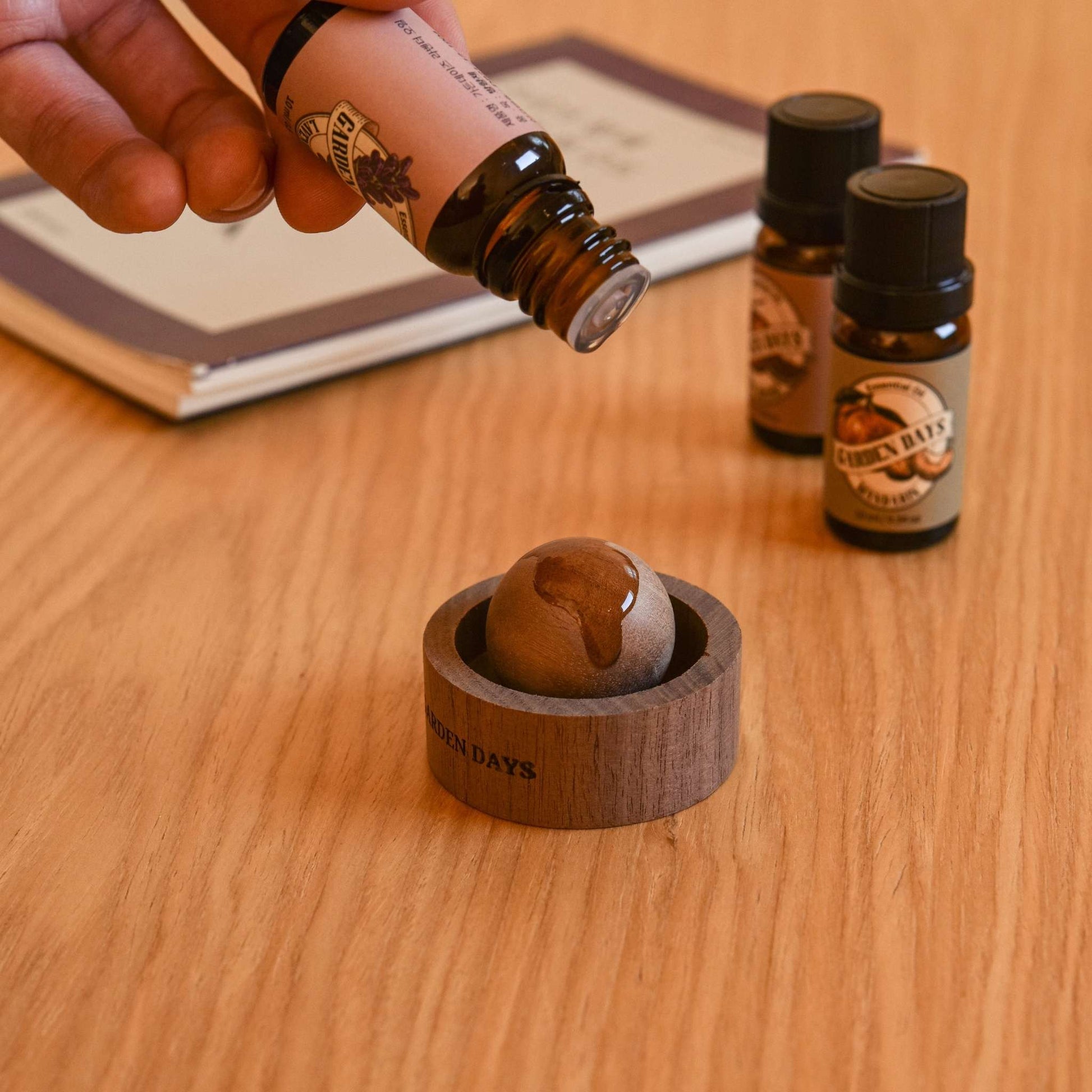 나무 책상 위에 올려둔 호두 브리즈볼에 에센셜오일을 떨어뜨리는 손과 옆에 세워진 가든데이즈 에센셜오일 병들이 보이는 사용 연출 사진