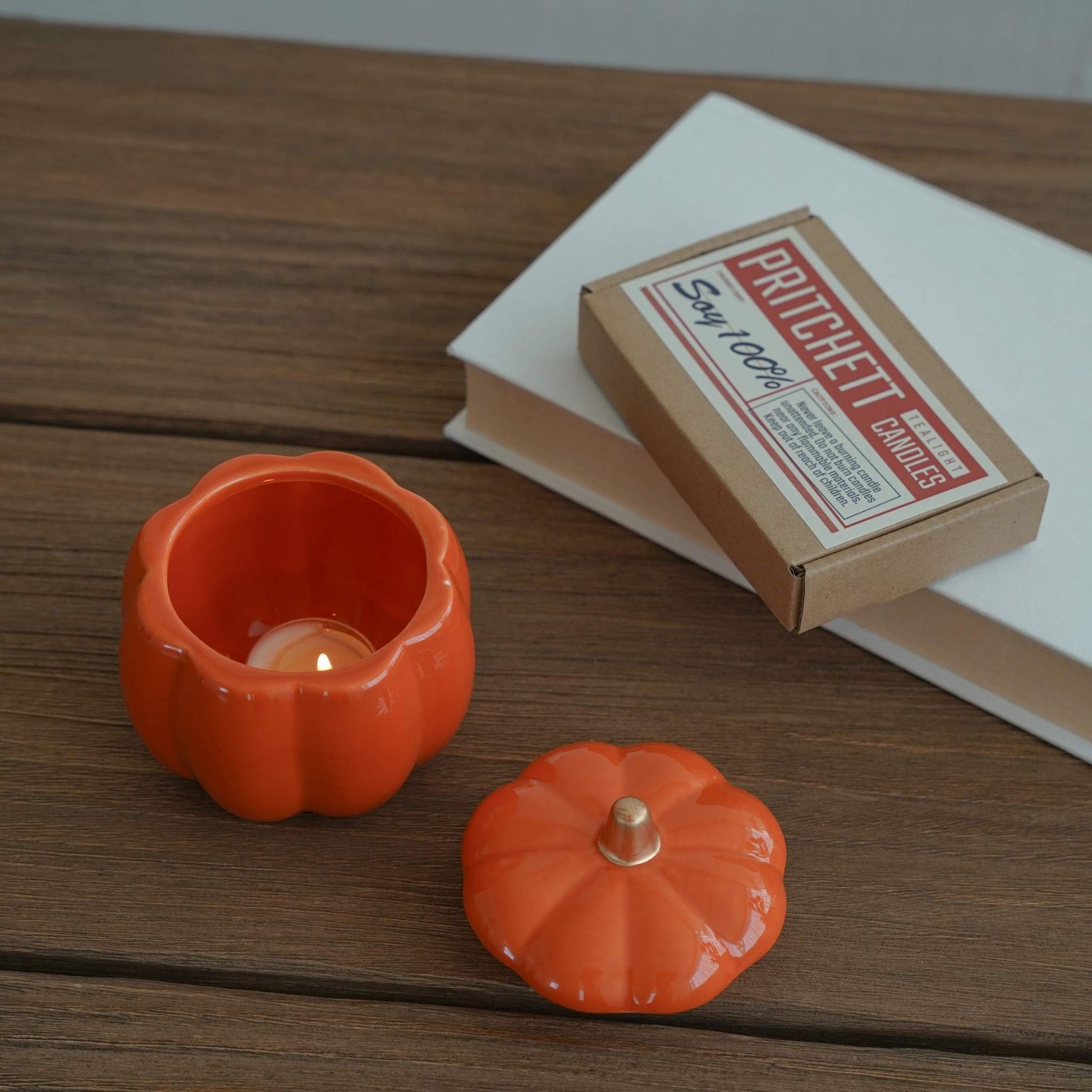 나무 책상 위 책과 캔들 옆에 놓인 주황색 호박 캔들 홀더와 뚜껑이 함께 보이는 가을 분위기 연출 사진