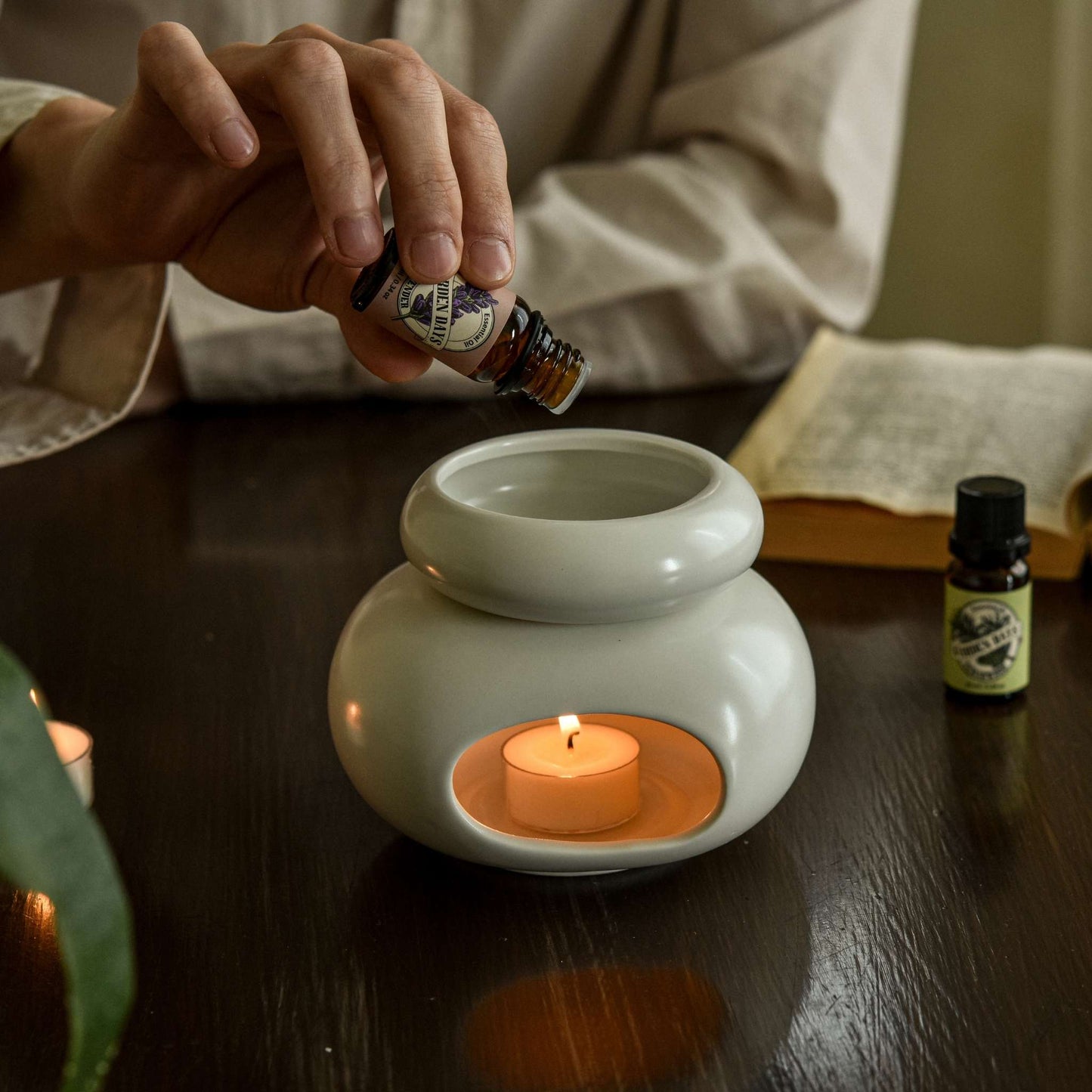테이블 위 화이트 조약돌 오일버너 상단에 에센셜오일을 떨어뜨리는 손과 옆에 놓인 가든데이즈 에센셜오일 병 두 개가 보이는 사용 연출 사진