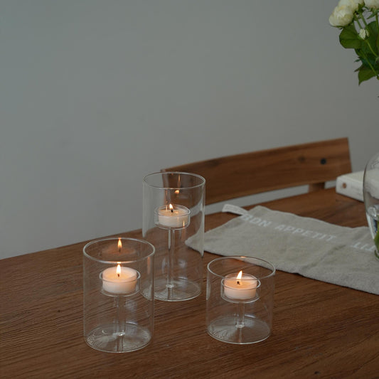 나무 테이블 위 서로 높이가 다른 투명 유리 캔들 홀더 세 개와 안에서 켜진 티라이트 캔들이 보이는 테이블 연출 사진