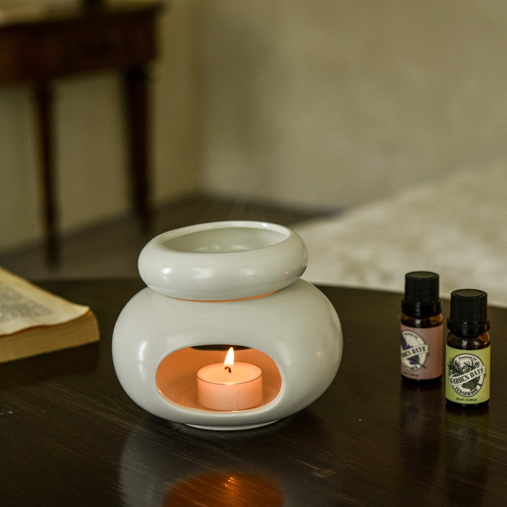 어두운 나무 테이블 위에 단독으로 놓인 화이트 조약돌 오일버너와 안에서 은은하게 빛나는 티라이트 캔들, 옆에 세워둔 가든데이즈 에센셜오일 병이 보이는 연출 사진
