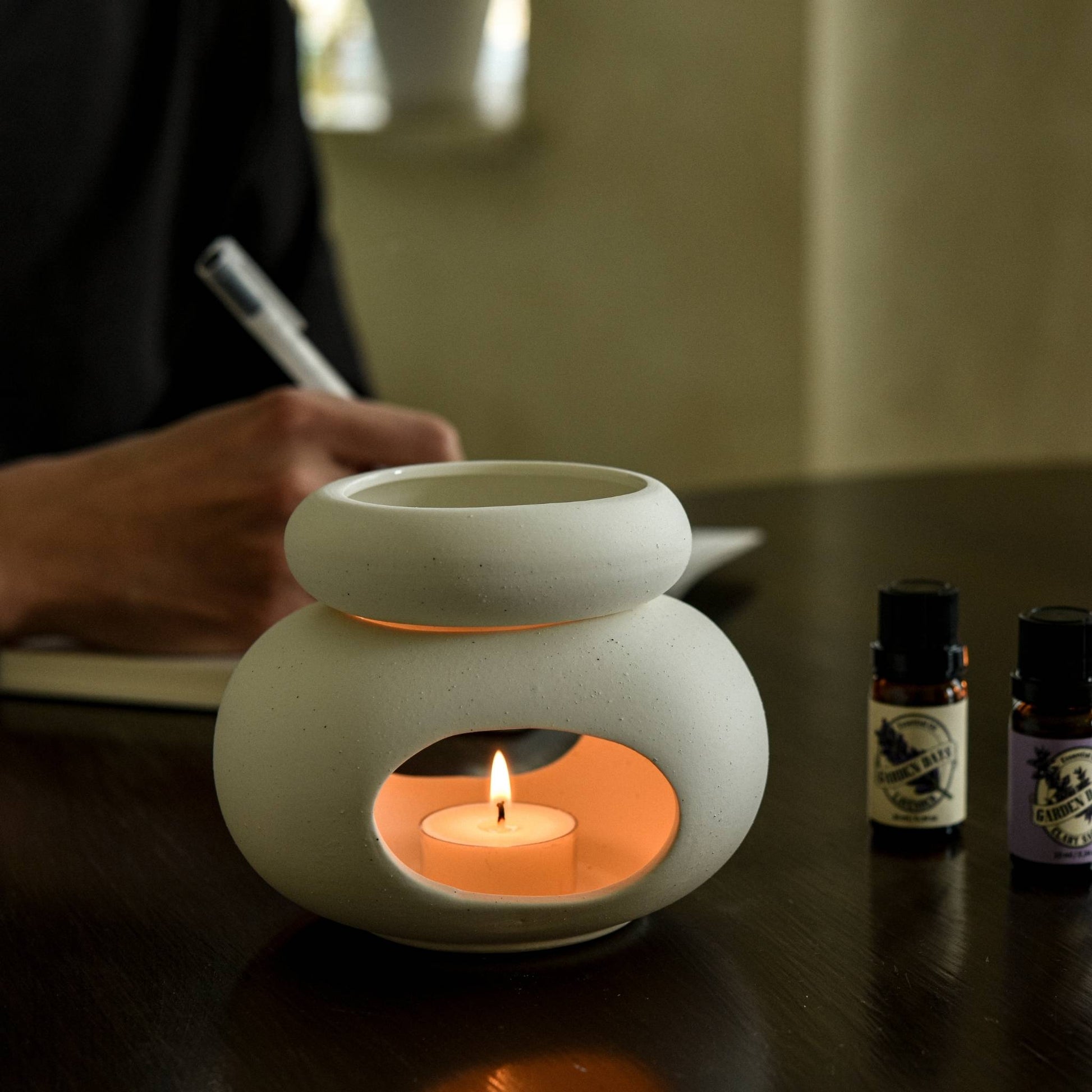 노트를 적고 있는 사람 앞 테이블 위에서 따뜻하게 불이 켜진 조약돌 오일버너와 가든데이즈 에센셜오일 병이 함께 놓인 조용한 작업 공간 연출 사진