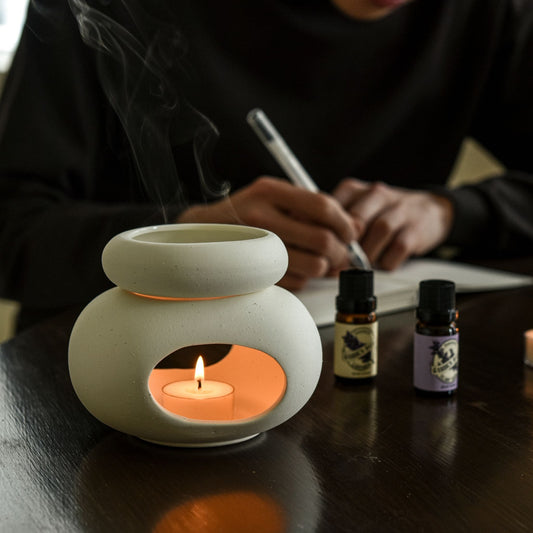 어두운 테이블 위에 놓인 오일버너와 불이 켜진 티라이트 캔들, 뒤로 노트를 적고 있는 사람의 손과 피어오르는 연기가 보이는 연출 사진