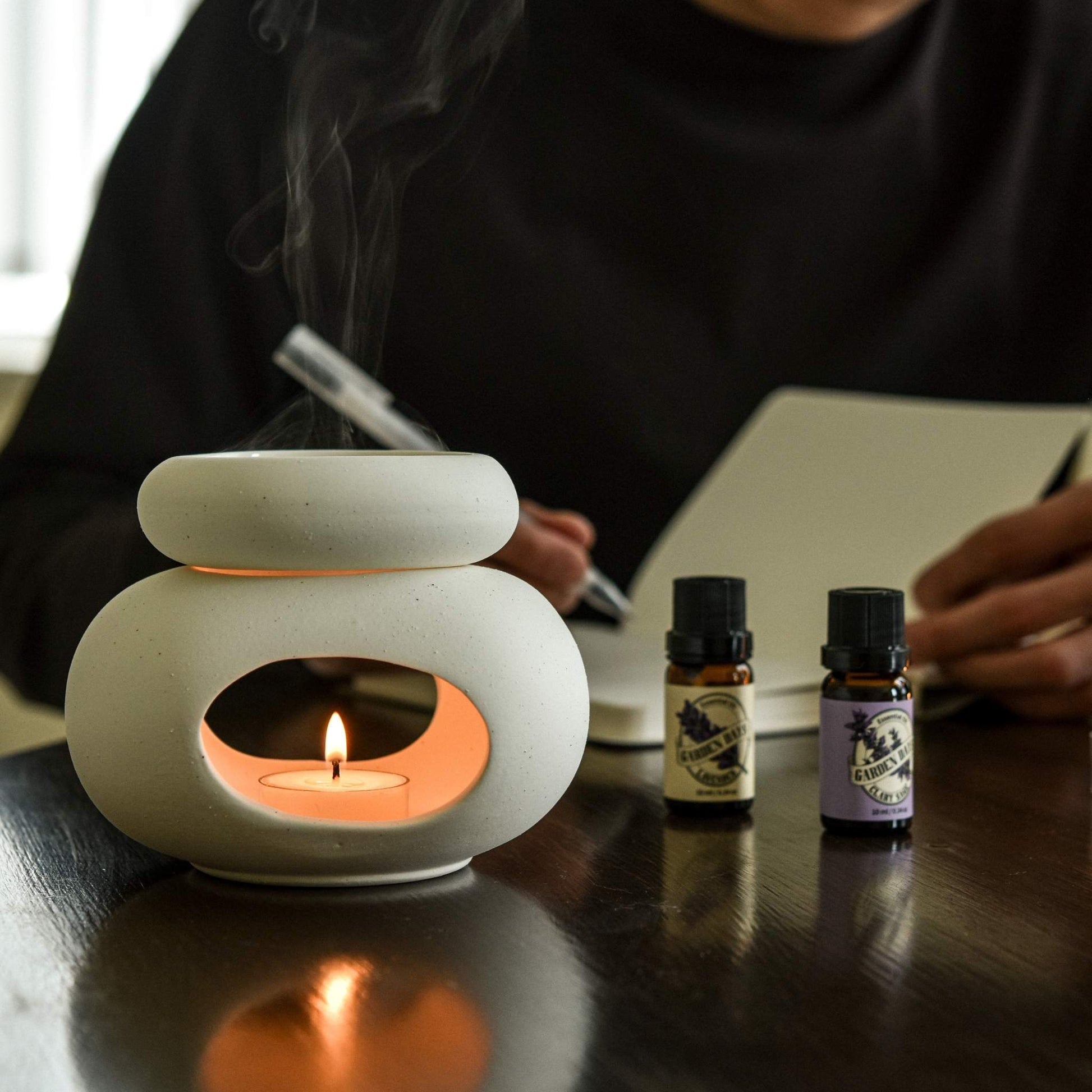 어두운 톤 책상 위에서 글을 쓰는 손 옆으로, 티라이트 캔들이 타고 있는 조약돌 오일버너와 가든데이즈 에센셜오일 3종이 함께 놓인 사용 연출 사진