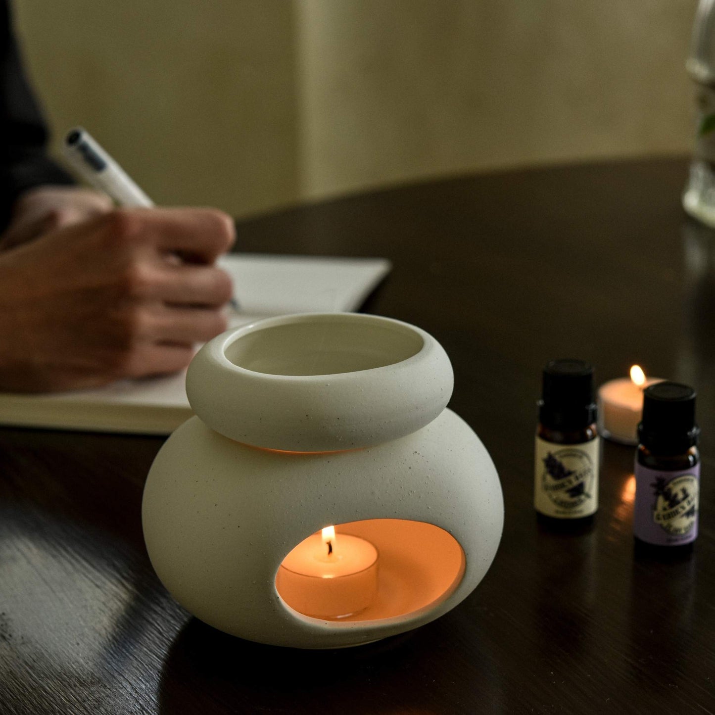 테이블 위 노트와 펜이 놓인 공간에서 조약돌 샌드 오일버너와 불이 켜진 티라이트 캔들, 가든데이즈 에센셜오일 병 두 개가 함께 보이는 작업 공간 연출 사진