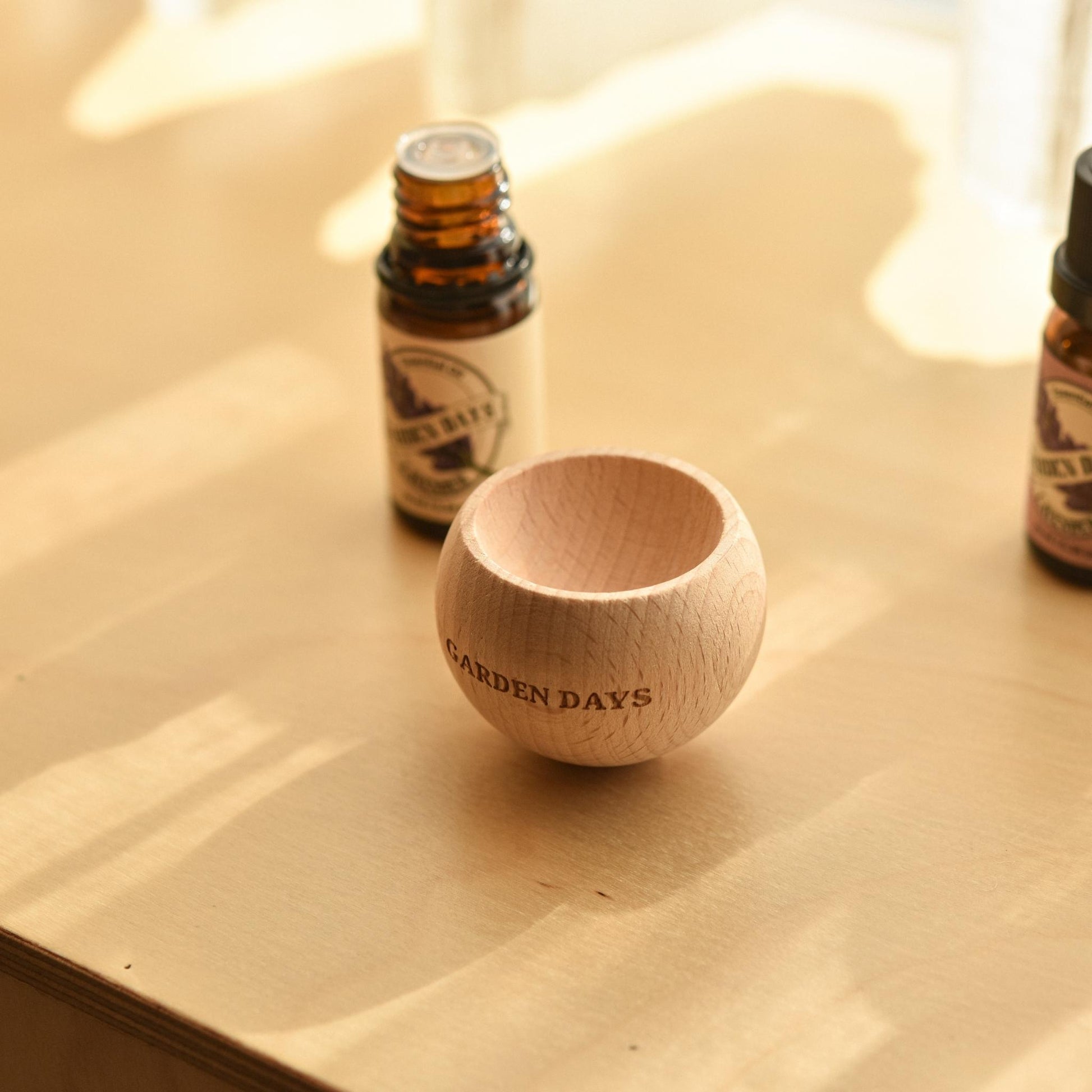 밝은 테이블 위에 놓인 가든데이즈 우드 브리즈볼과 뒤쪽에 세워진 에센셜오일 병이 보이는 제품 연출 사진