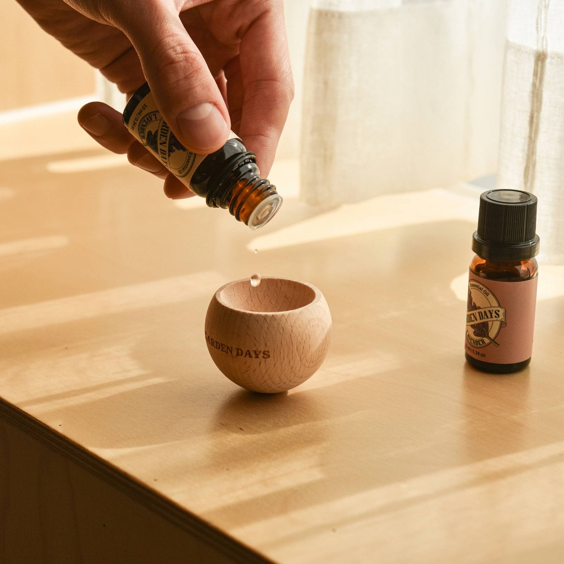 창가 쪽 나무 테이블 위에 놓인 가든데이즈 우드 브리즈볼 상단에 에센셜오일을 떨어뜨리는 손과 옆에 선 오일 병이 보이는 사용 연출 사진