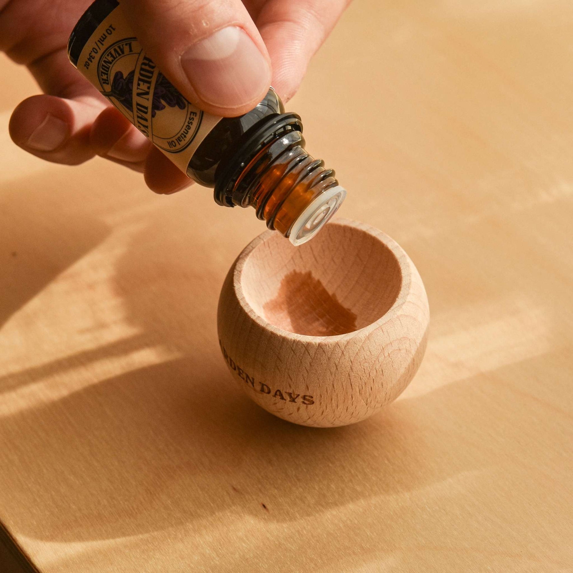 나무 표면 위에 올려진 가든데이즈 우드 브리즈볼에 에센셜오일을 가까이에서 떨어뜨리는 손이 보이는 연출 사진