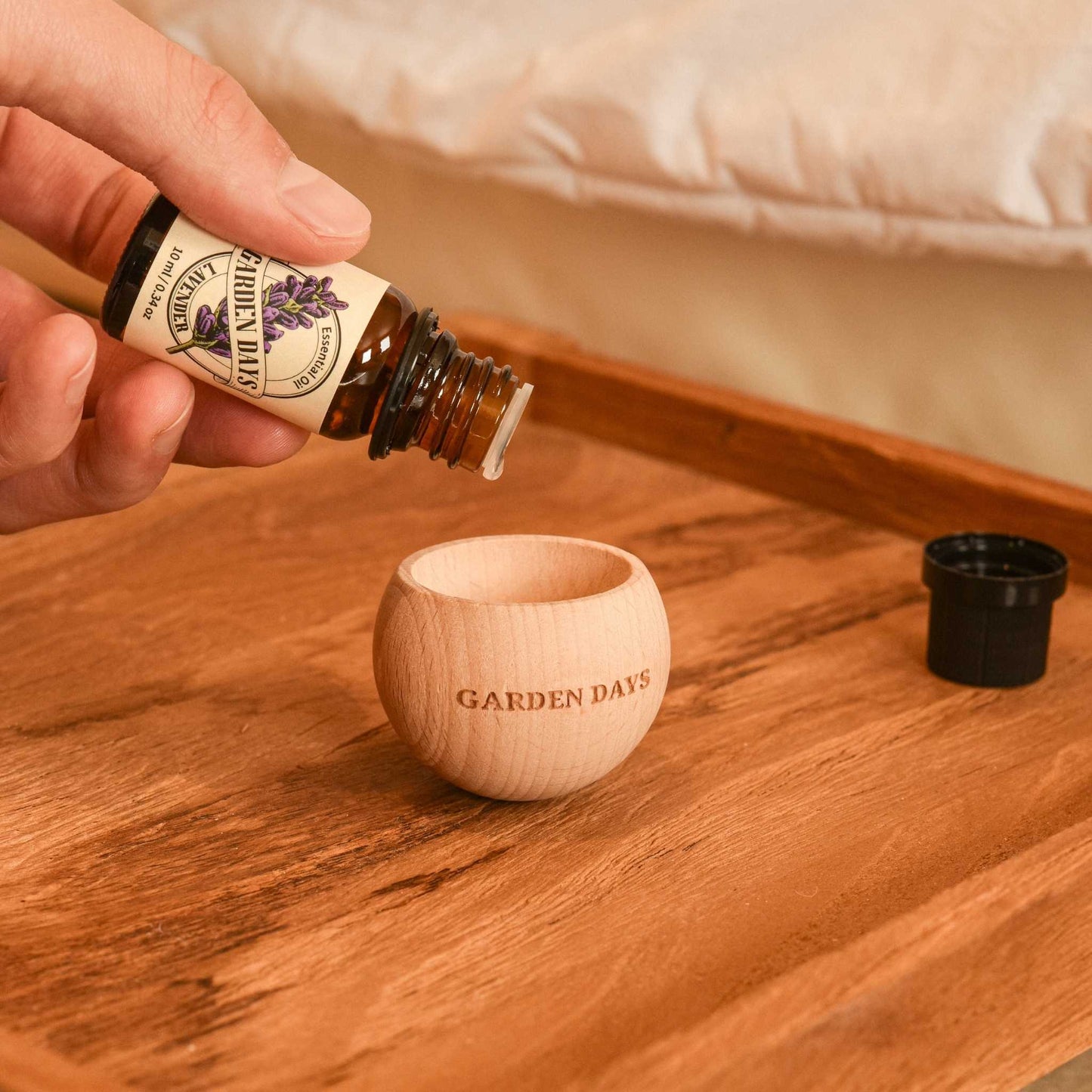침대 옆 나무 테이블 위 가든데이즈 우드 브리즈볼과 가든데이즈 에센셜오일을 떨어뜨리는 손이 보이는 연출 사진
