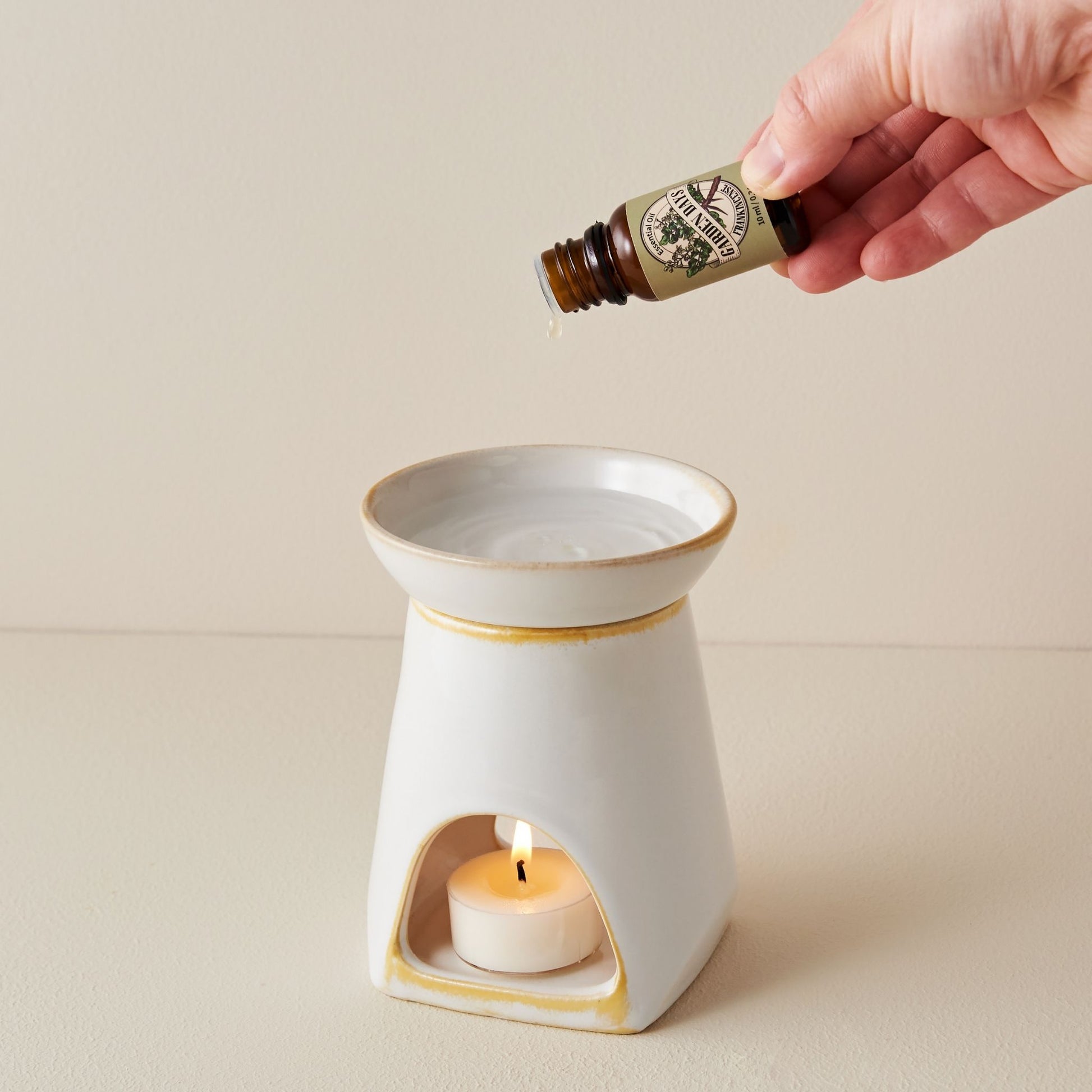 크림색 배경 위에 노란 가장자리가 포인트인 화이트 세라믹 옐로우 오일버너와 안에서 불이 켜진 티라이트 캔들, 옆에 놓인 노란 꽃 장식이 보이는 제품 연출 사진