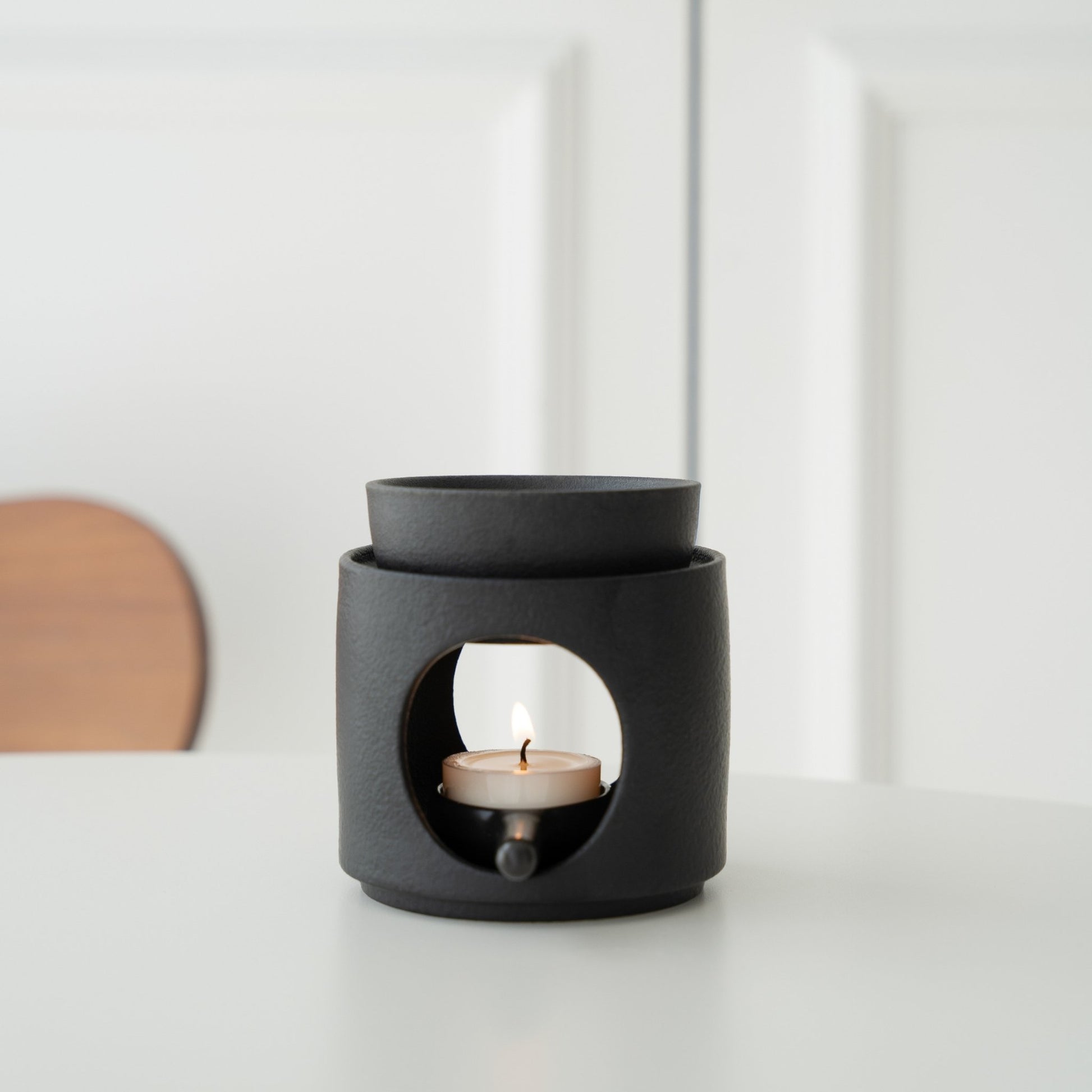 화이트 테이블 위에 단독으로 놓인 블랙 세라믹 오일버너와 안에서 켜진 티라이트 캔들 정면 사진