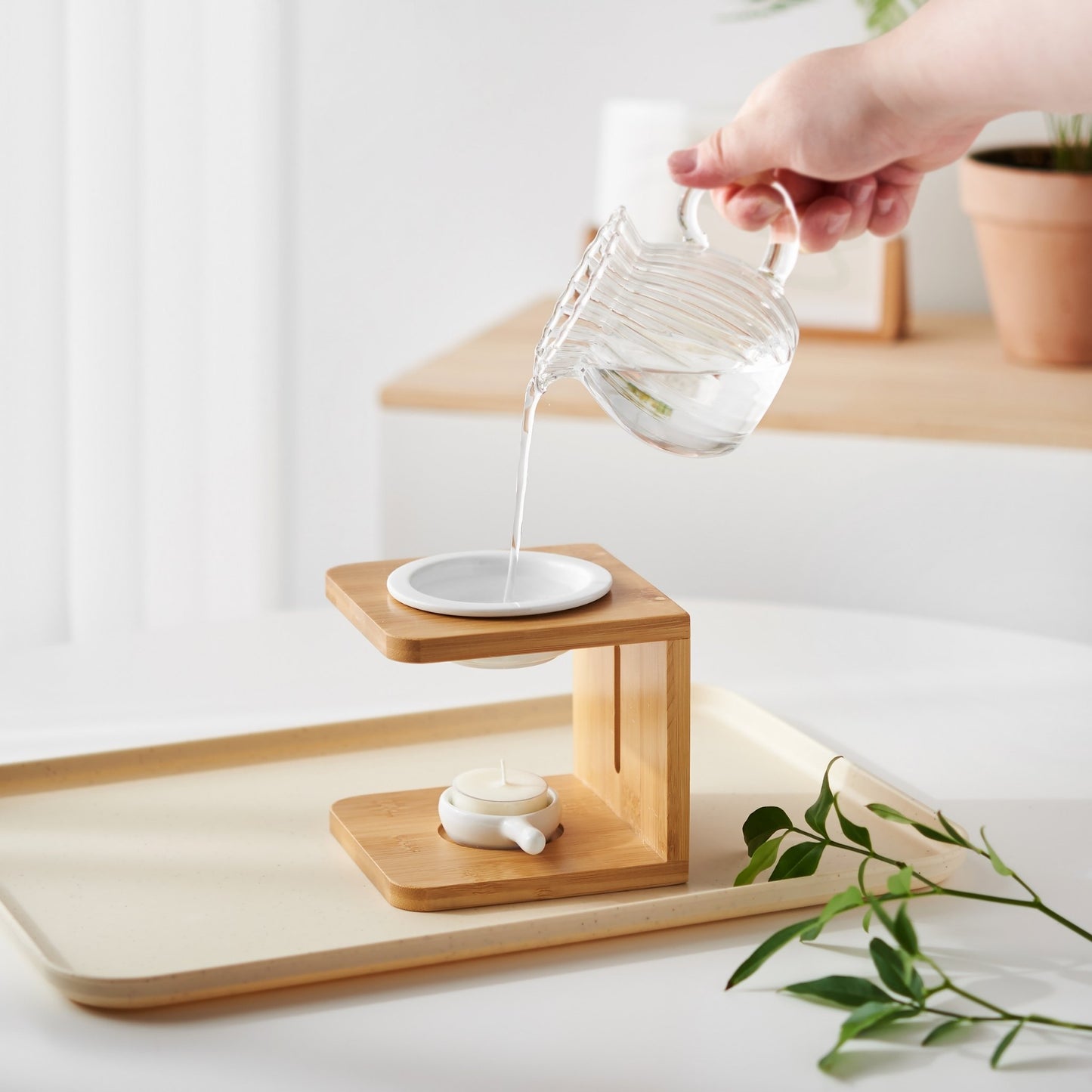 밝은 트레이 위 우드 오일버너 상단 물에 에센셜오일을 떨어뜨리는 손과 옆에 놓인 티라이트 캔들, 그린 잎 장식이 보이는 사용 연출 사진