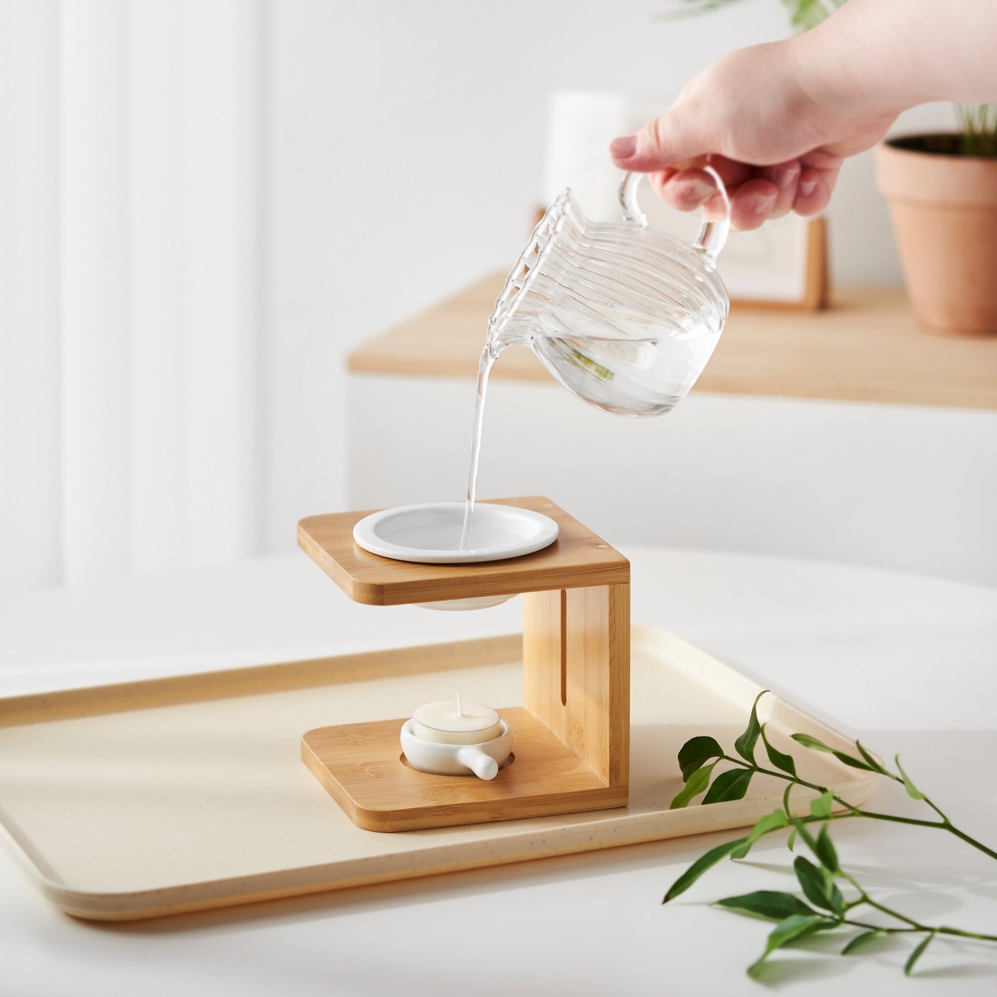 밝은 트레이 위 우드 오일버너 상단 물에 에센셜오일을 떨어뜨리는 손과 옆에 놓인 티라이트 캔들, 그린 잎 장식이 보이는 사용 연출 사진