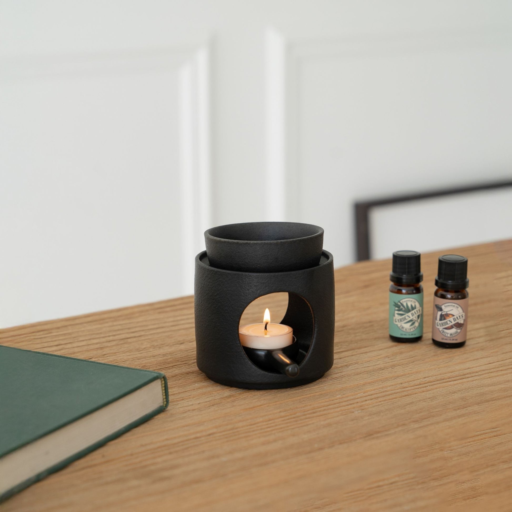 나무 테이블 위 노트와 가든데이즈 에센셜오일 병 두 개 옆에 놓인 블랙 세라믹 오일버너와 불이 켜진 티라이트 캔들이 보이는 연출 사진