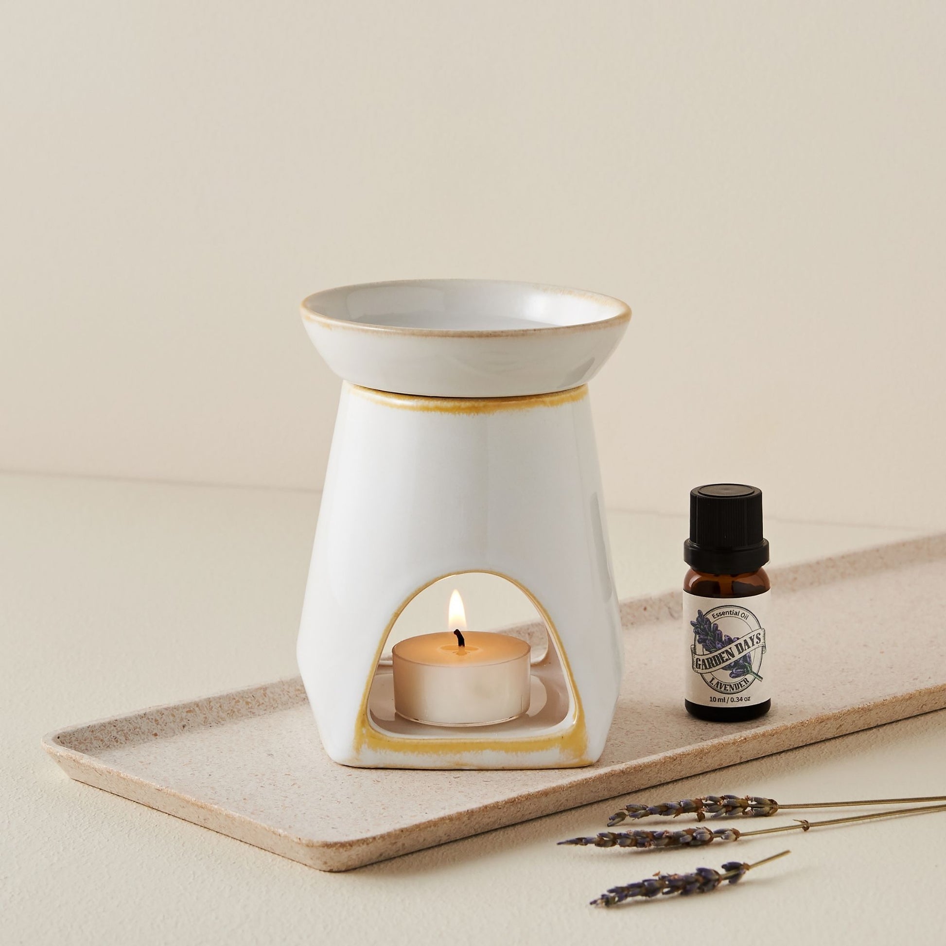 크림색 배경 위 직사각 트레이에 놓인 화이트 세라믹 옐로우 오일버너와 불이 켜진 티라이트 캔들, 옆에 놓인 가든데이즈 에센셜오일 병과 드라이 라벤더 스틱이 함께 연출된 사진
