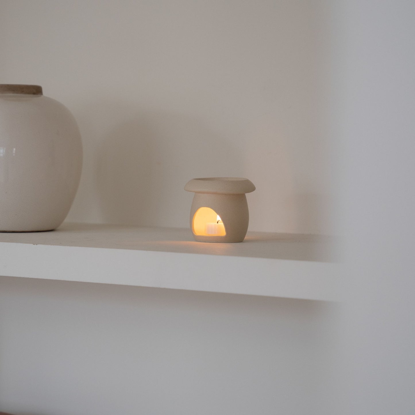 `화이트 선반 위 큰 화병 옆에 놓인 베이지 색 머쉬룸 오일버너와 안에서 켜진 티라이트 캔들 연출 사진`
