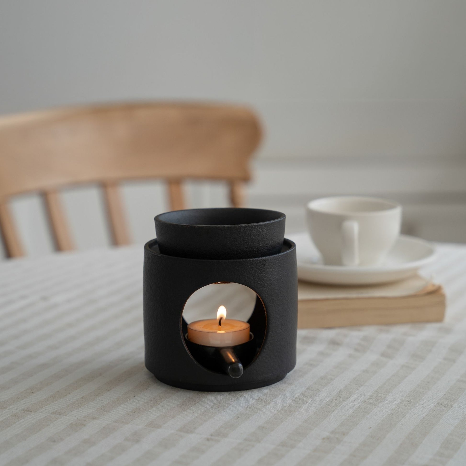 줄무늬 테이블보와 의자, 찻잔이 보이는 식탁 위에 놓인 블랙 세라믹 오일버너와 불이 켜진 티라이트 캔들 사진