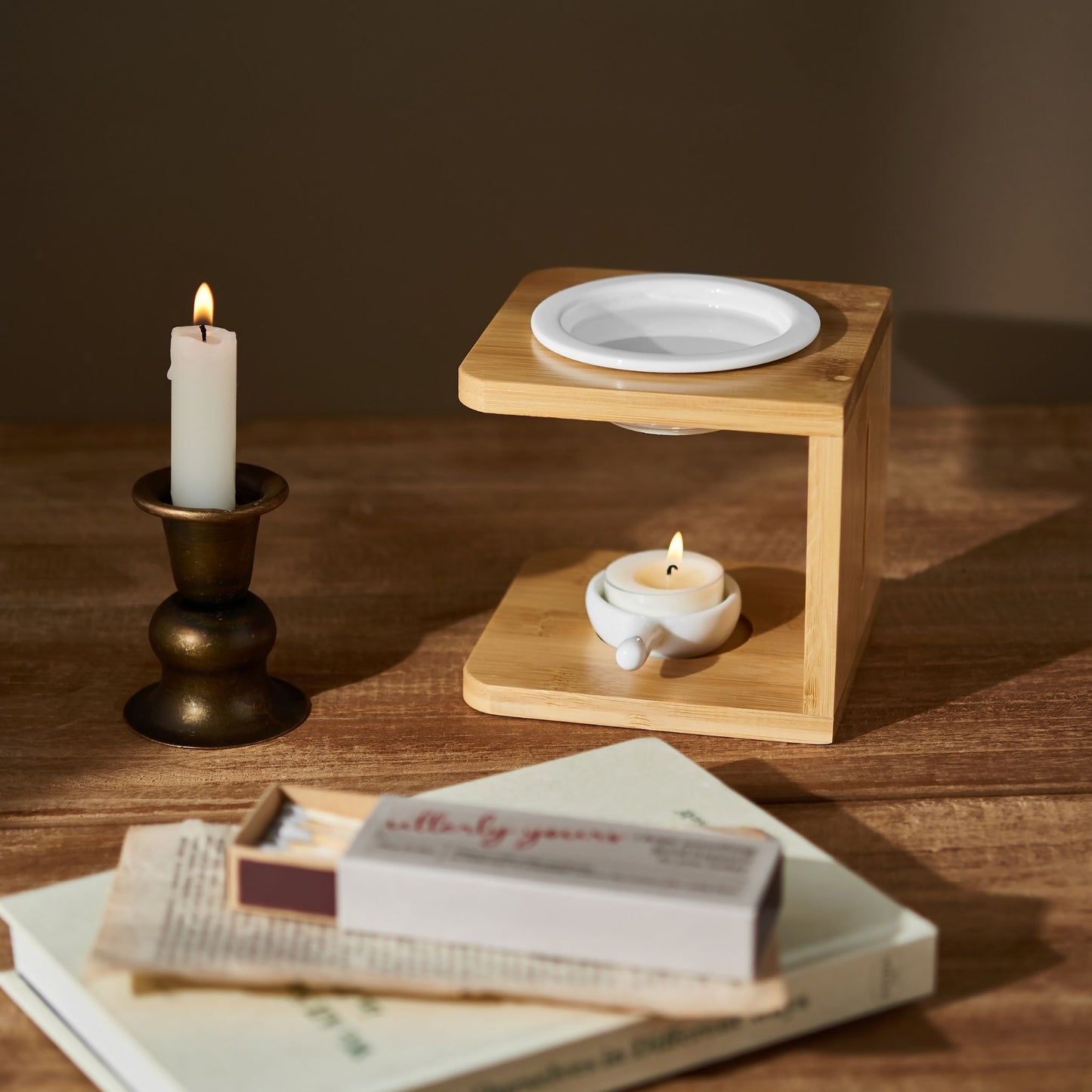 나무 테이블 위 촛대와 기둥 캔들, 우드 오일버너와 불이 켜진 티라이트 캔들, 앞쪽에 놓인 책과 성냥갑이 보이는 연출 사진
