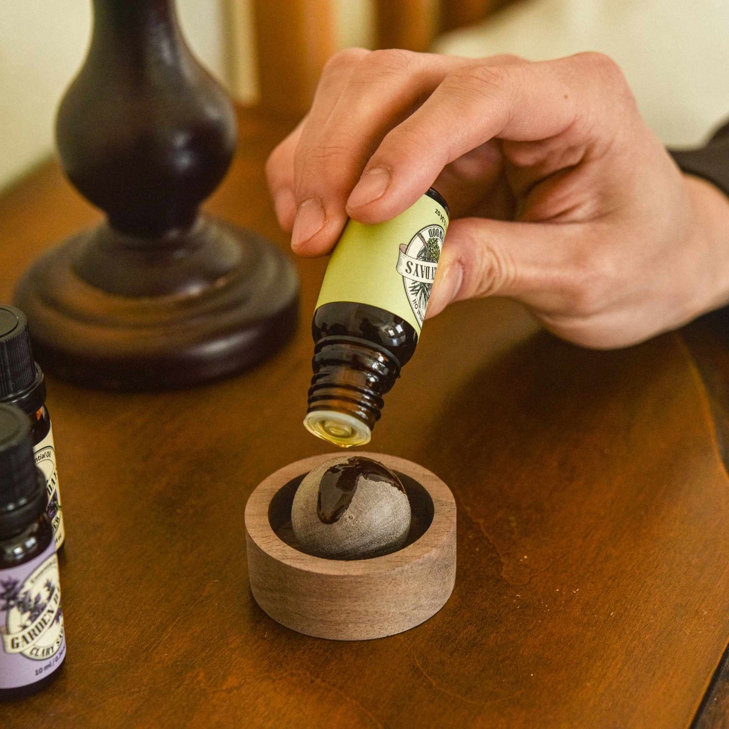 어두운 톤 테이블 위 호두 브리즈볼 위로 에센셜오일을 떨어뜨리는 손과 뒤쪽 스탠드, 가든데이즈 에센셜오일 병이 함께 보이는 사용 연출 사진