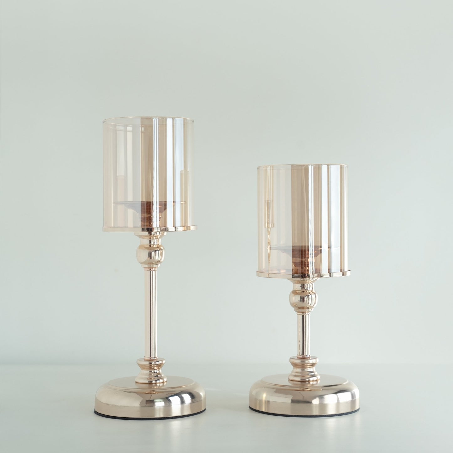 밝은 배경 앞에 골드 베이스의 유리 갓이 있는 스탠드 캔들 홀더 두 개를 나란히 세운 제품 단독 사진