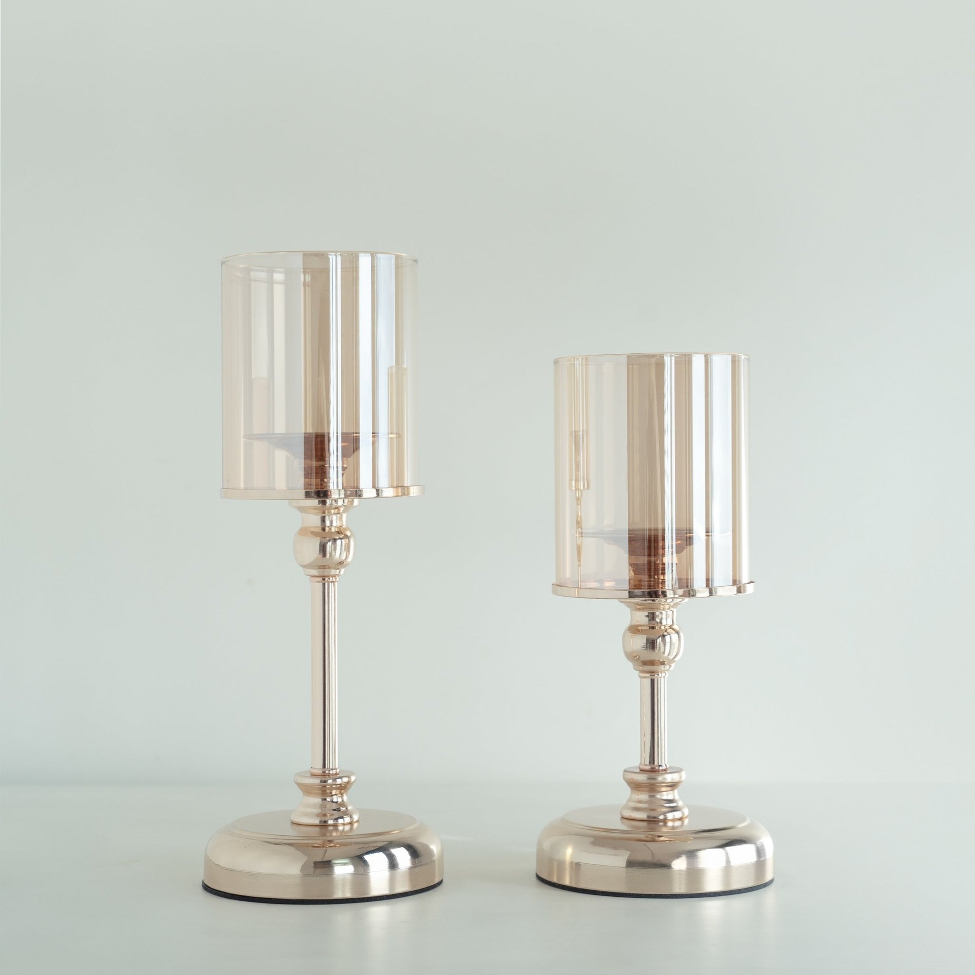 밝은 배경 앞에 골드 베이스의 유리 갓이 있는 스탠드 캔들 홀더 두 개를 나란히 세운 제품 단독 사진