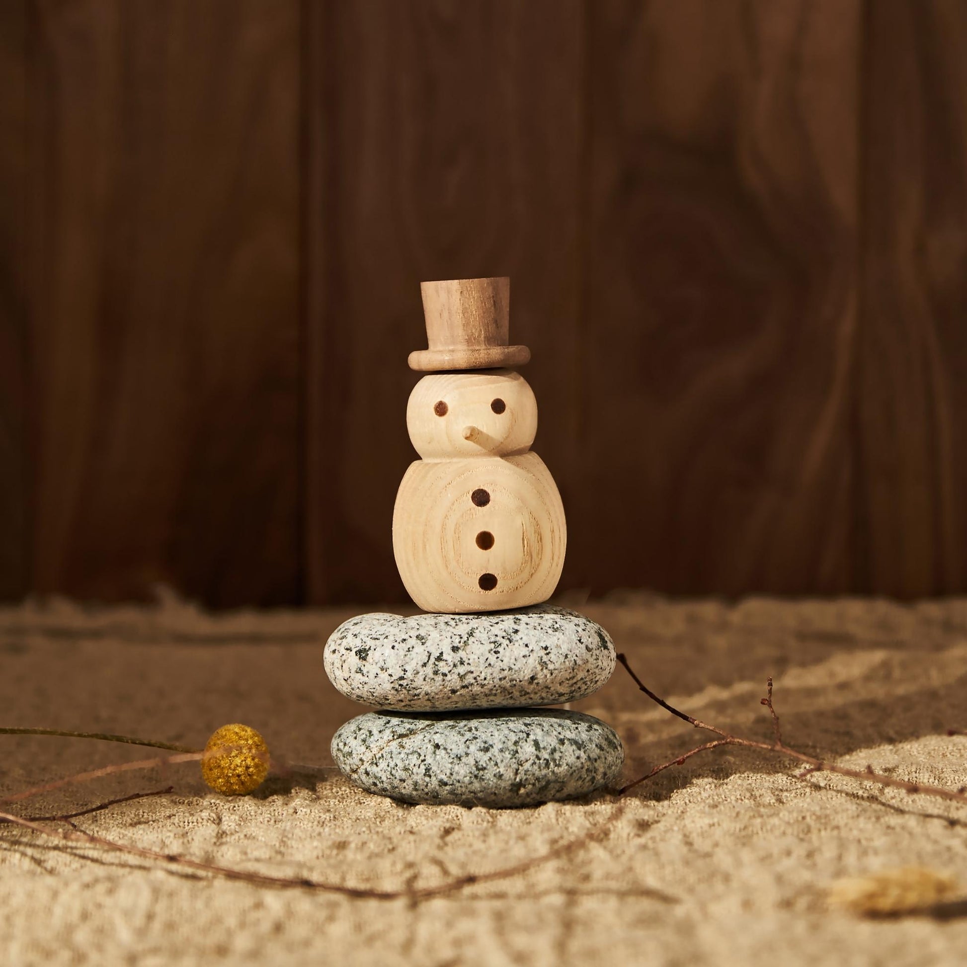 따뜻한 조명 아래 조약돌 위에 올려진 우드 스노우맨 디퓨저와 옆의 작은 드라이 플라워가 보이는 클로즈업 사진