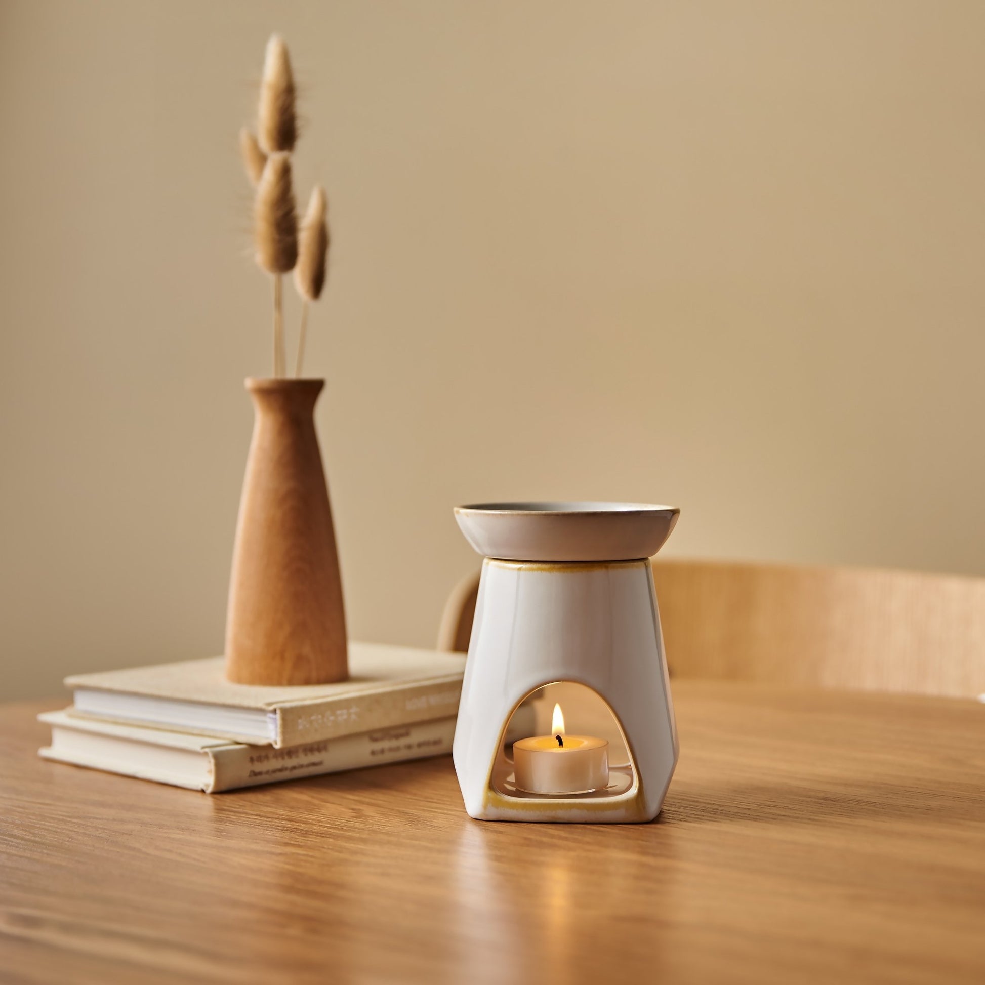 나무 테이블 위 책 더미와 우드 꽃병, 성냥갑 사이에 놓인 화이트 세라믹 옐로우 오일버너와 불이 켜진 티라이트 캔들이 보이는 따뜻한 분위기의 연출 사진