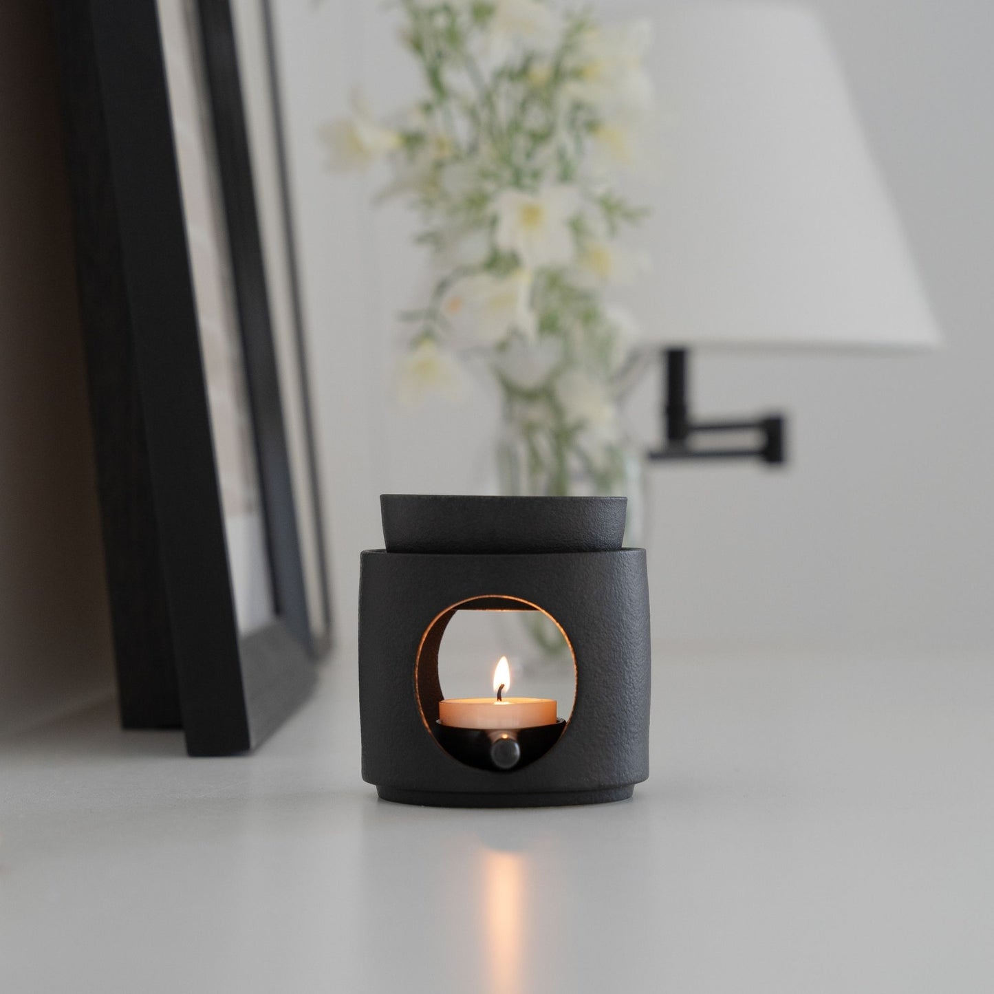 화병과 액자 옆에 놓인 블랙 세라믹 오일버너와 불이 켜진 티라이트 캔들이 보이는 미니멀 인테리어 연출 사진
