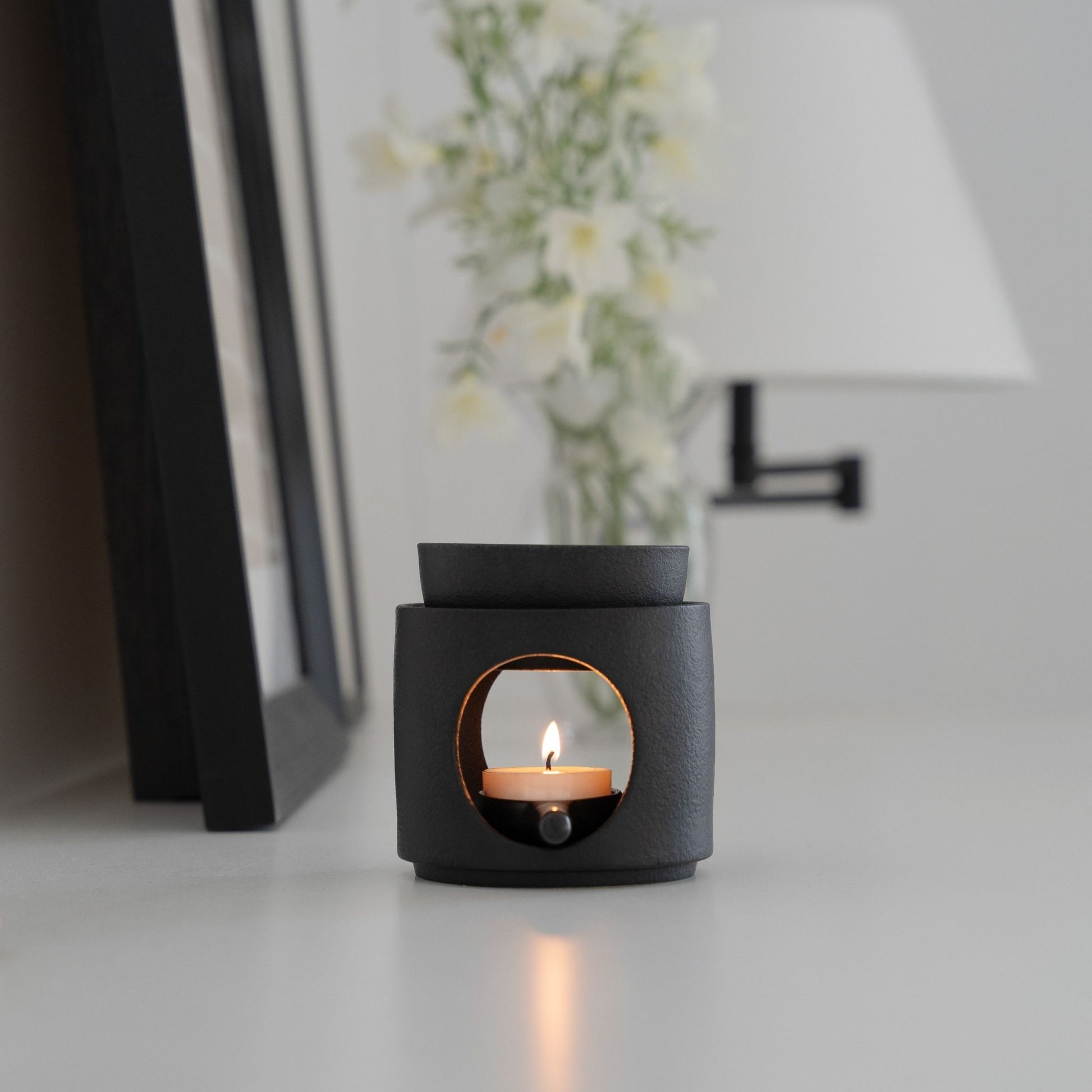 화병과 액자 옆에 놓인 블랙 세라믹 오일버너와 불이 켜진 티라이트 캔들이 보이는 미니멀 인테리어 연출 사진