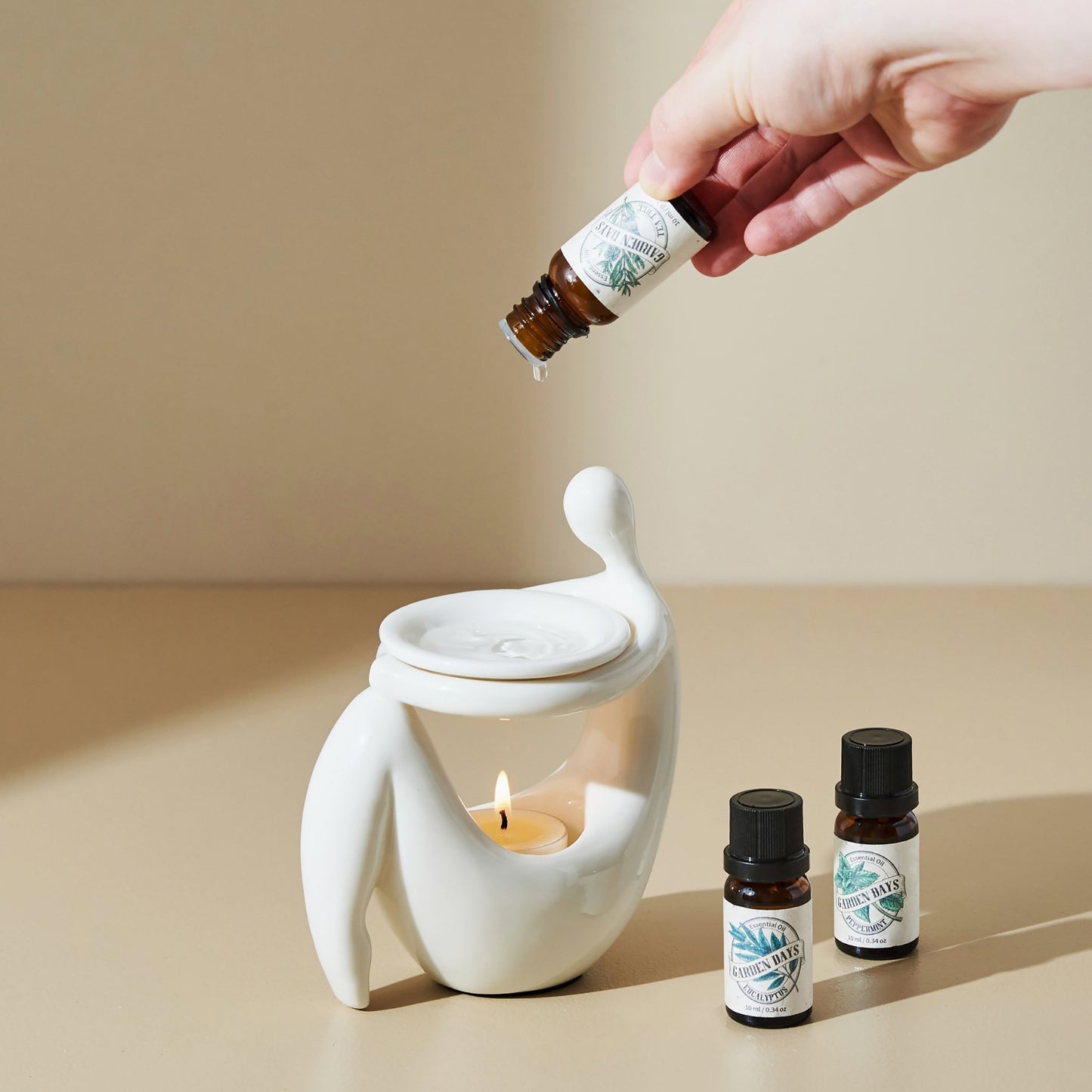 베이지 톤 배경 위 화이트 조형적인 세라믹 오일버너 상단에 에센셜오일을 떨어뜨리는 손과, 앞에 놓인 가든데이즈 에센셜오일 병 두 개, 안에서 켜진 티라이트 캔들이 보이는 사용 연출 사진