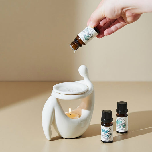 베이지 톤 배경 위 화이트 조형적인 세라믹 오일버너 상단에 에센셜오일을 떨어뜨리는 손과, 앞에 놓인 가든데이즈 에센셜오일 병 두 개, 안에서 켜진 티라이트 캔들이 보이는 사용 연출 사진