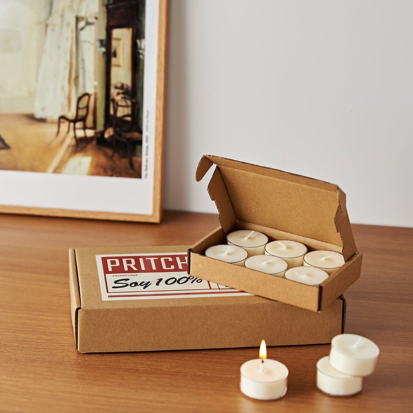 액자 앞 나무 테이블 위에 놓인 소이 티라이트 캔들 6개입 작은 상자와 33개입 큰 상자, 상자 앞에 꺼내 둔 티라이트 캔들 몇 개가 보이는 메인 제품 사진