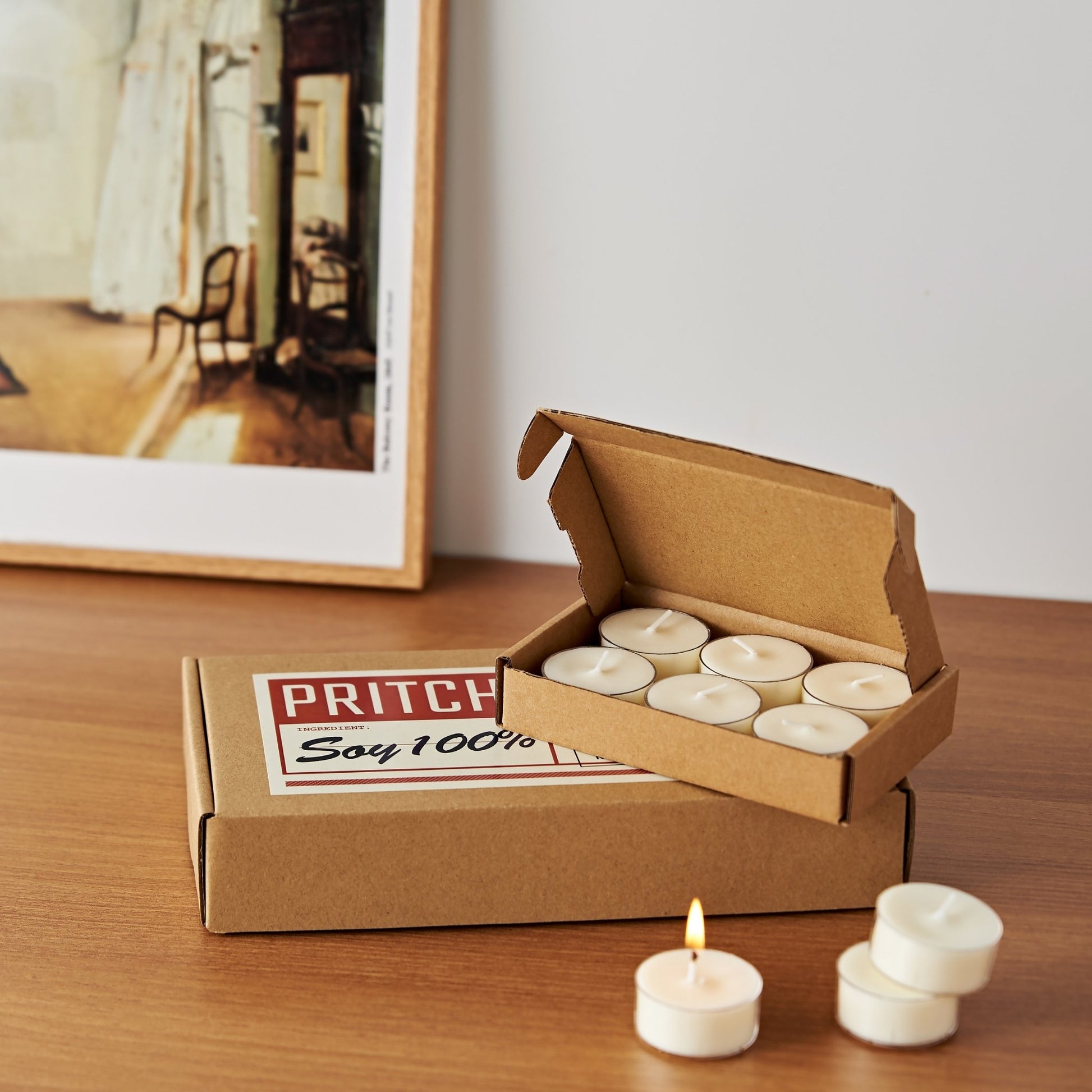 액자 앞 나무 테이블 위에 놓인 소이 티라이트 캔들 6개입 작은 상자와 33개입 큰 상자, 상자 앞에 꺼내 둔 티라이트 캔들 몇 개가 보이는 메인 제품 사진