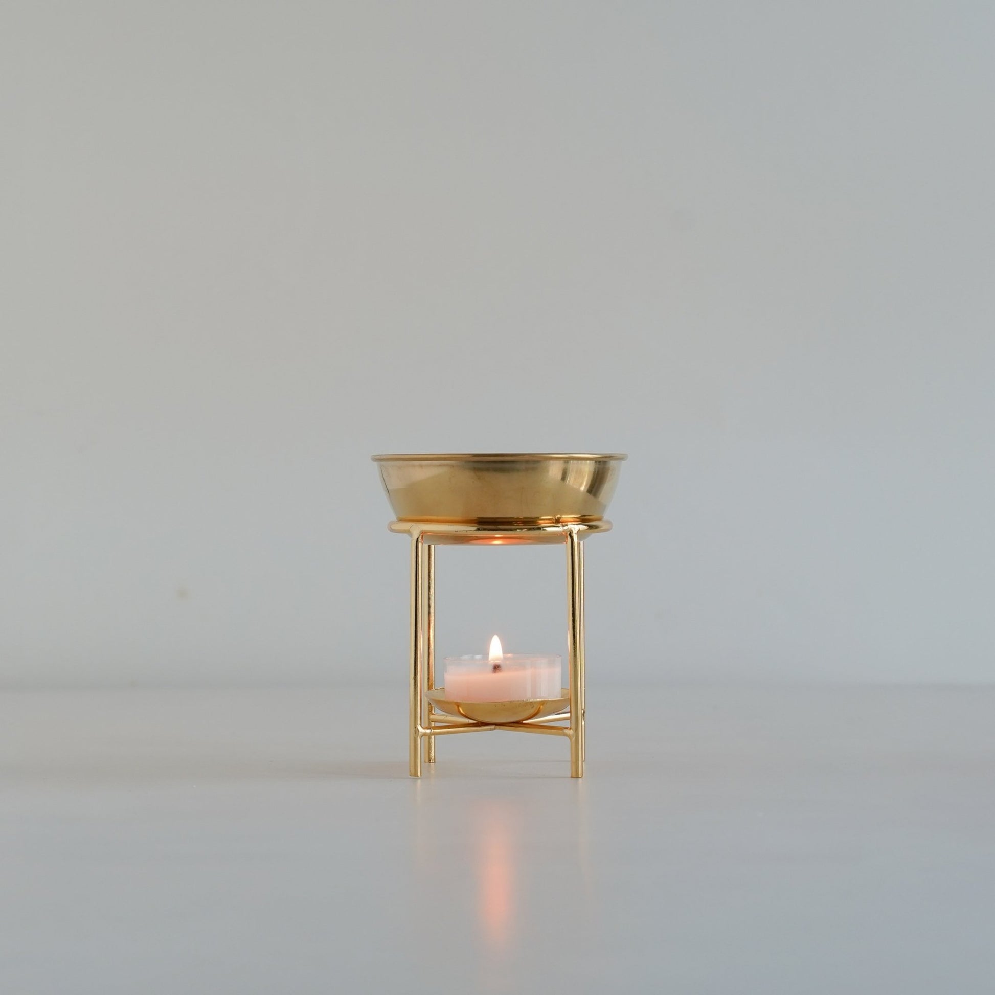 연한 배경 앞 단독으로 놓인 골드 스탠드 오일버너와 안에서 은은하게 빛나는 티라이트 캔들을 정면에서 담은 제품 사진
