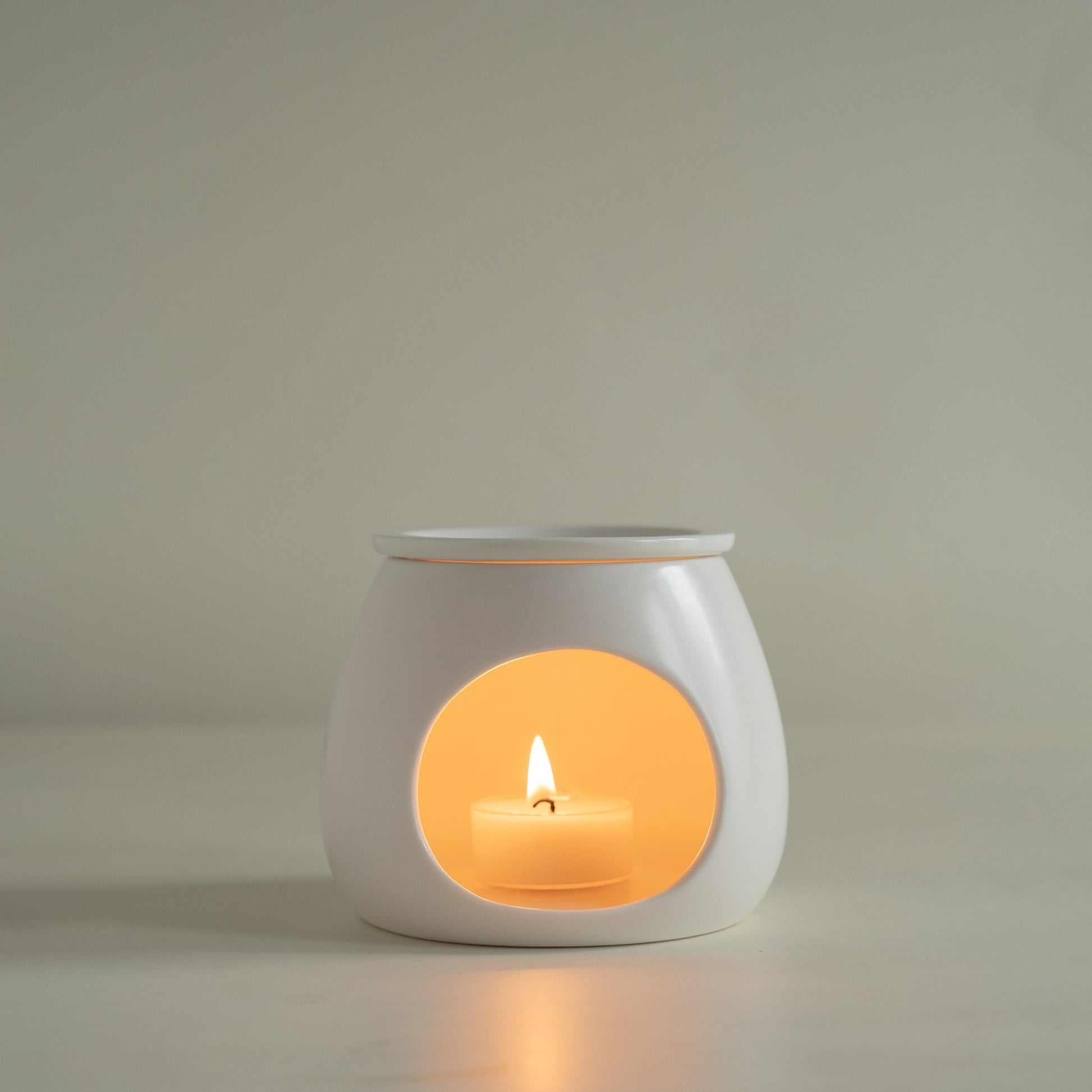 연한 배경 앞에 단독으로 놓인 화이트 세라믹 오일버너와 안에서 따뜻하게 빛나는 티라이트 캔들이 보이는 정면 제품 사진