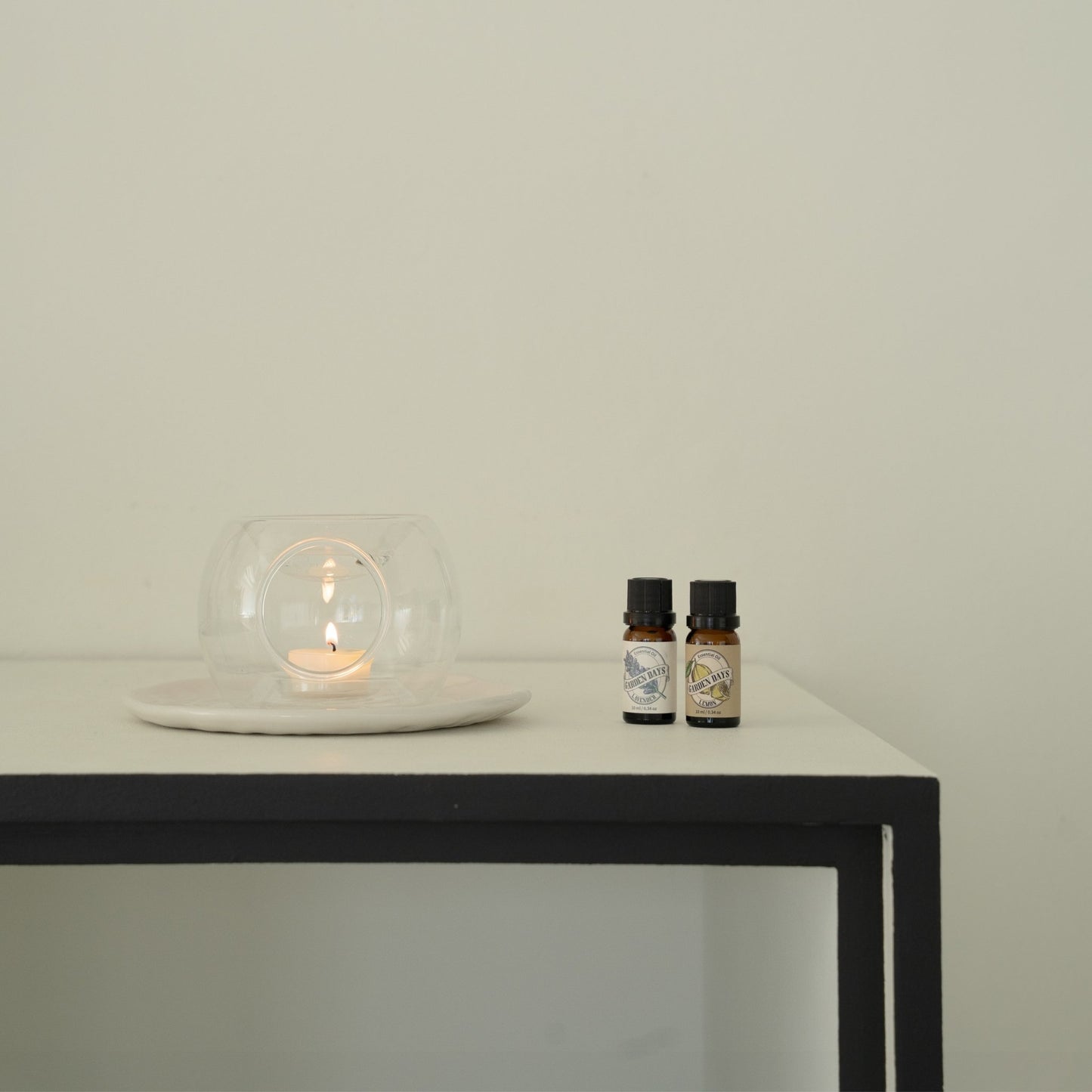 블랙 프레임 테이블 위 투명 유리 티라이트 홀더와 불이 켜진 티라이트 캔들, 옆에 세워둔 가든데이즈 에센셜오일 병 두 개가 보이는 연출 사진