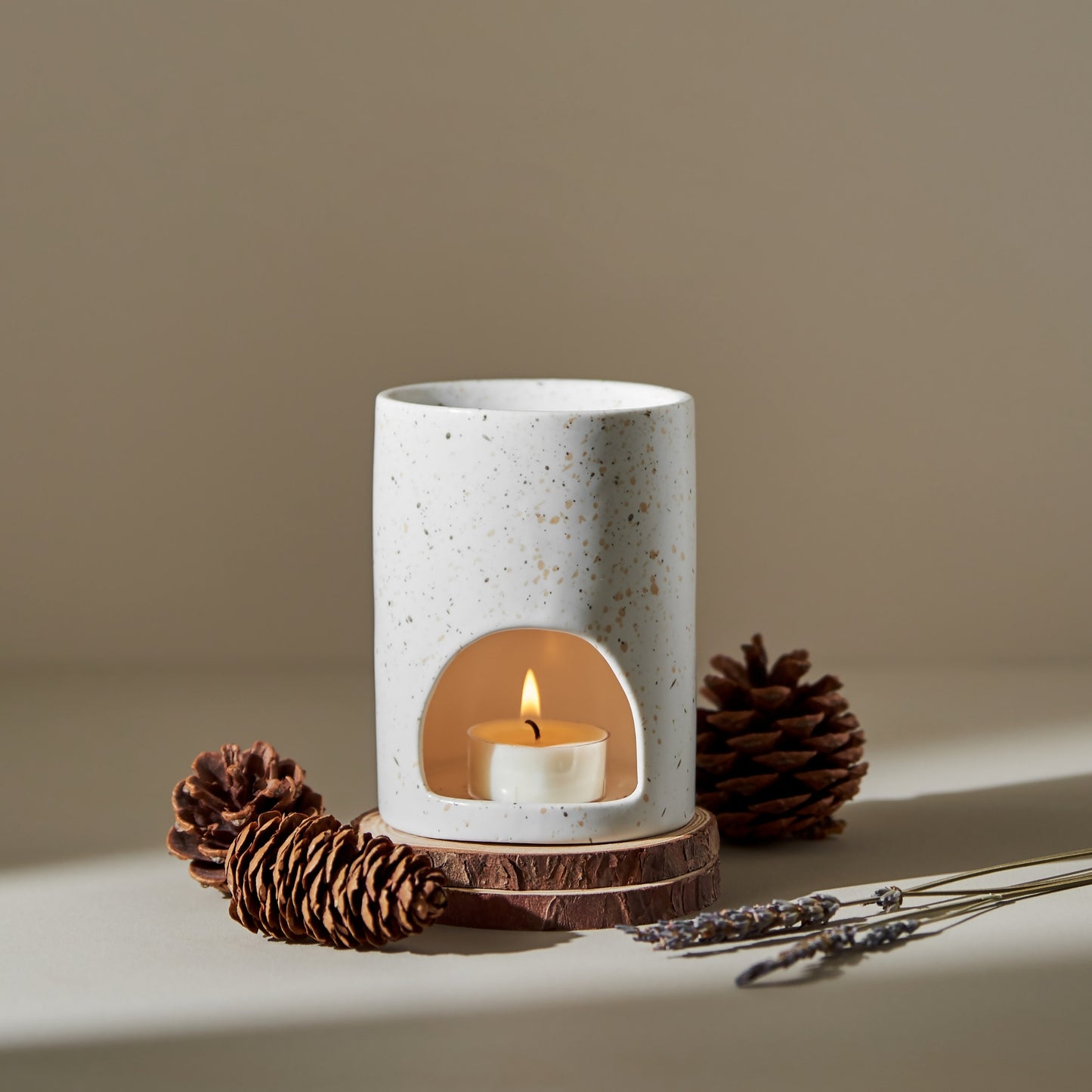 크림색 배경 위 나무 조각과 솔방울, 드라이 라벤더 위에 놓인 마블 텍스처 오일버너와 안에서 은은하게 켜진 티라이트 캔들이 보이는 겨울 느낌 연출 사진